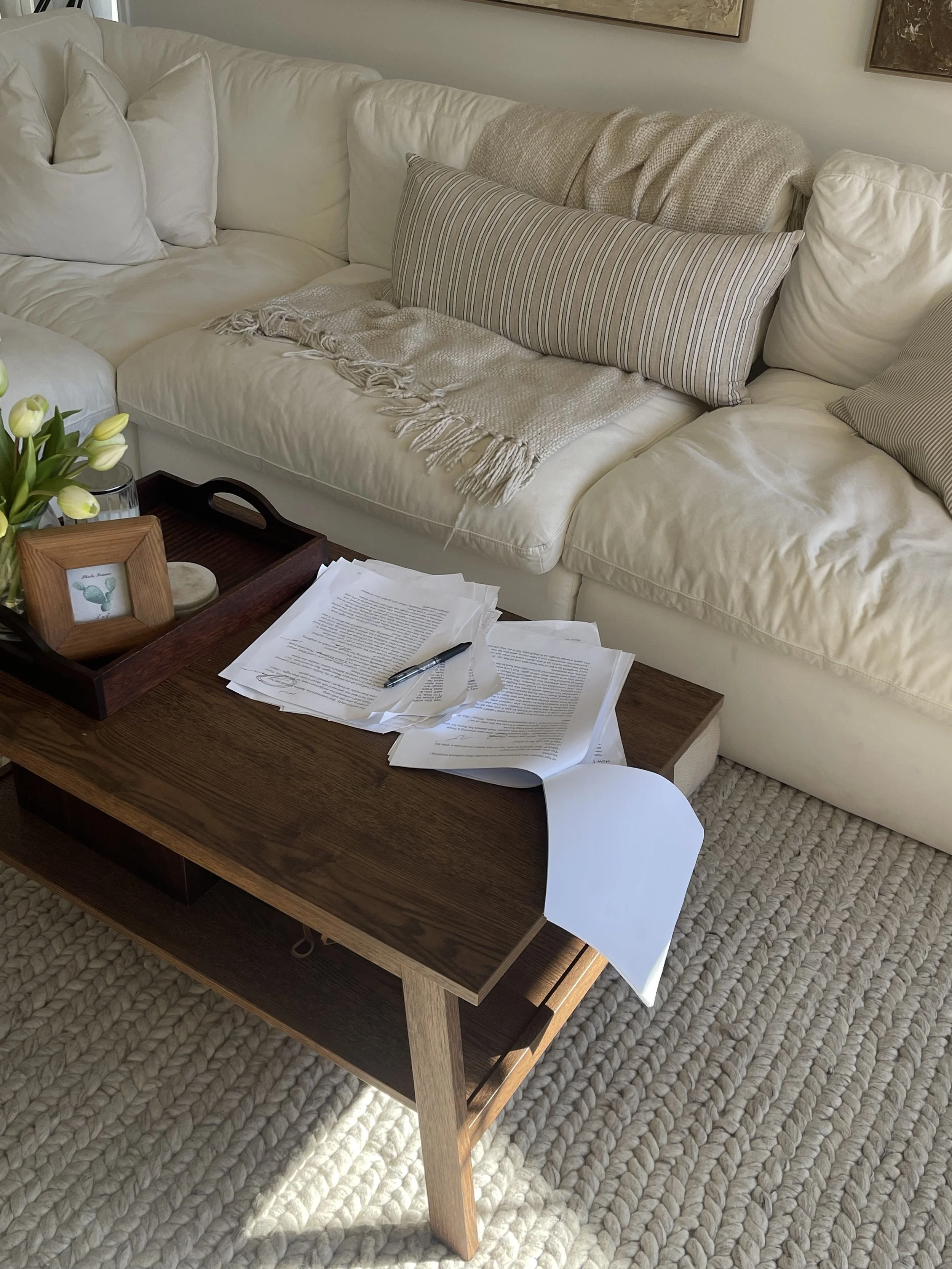 Living room with white sectional sofa, beige and striped throw pillows, a beige blanket, a wooden coffee table with papers, a pen, a glass vase with white tulips, and framed artwork on the wall. The room has a textured beige rug on the floor.