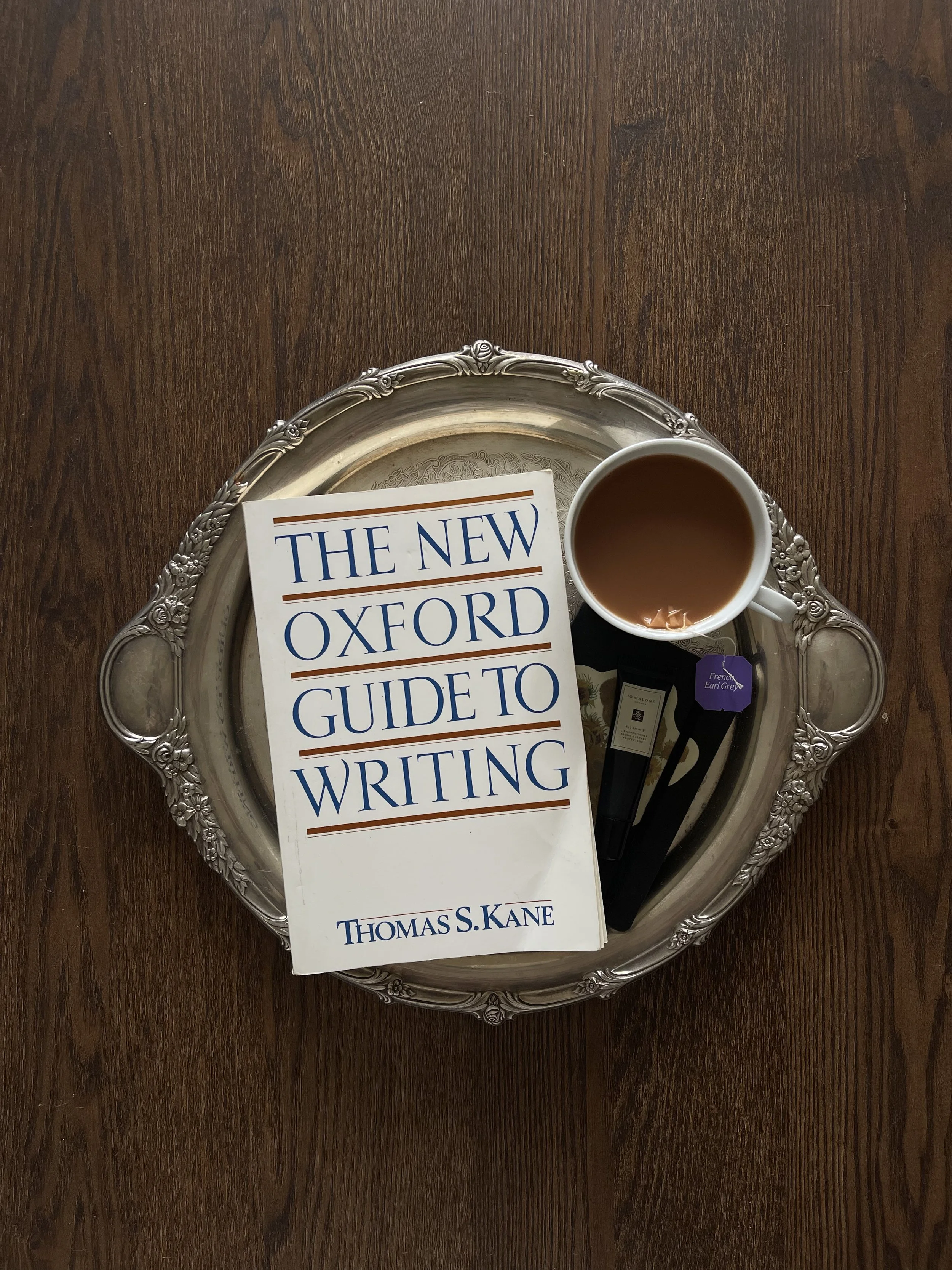 A silver tray holding a book titled 'The New Oxford Guide to Writing' by Thomas S. Kane, a cup of coffee, and some tea bags, all placed on a dark wooden surface.