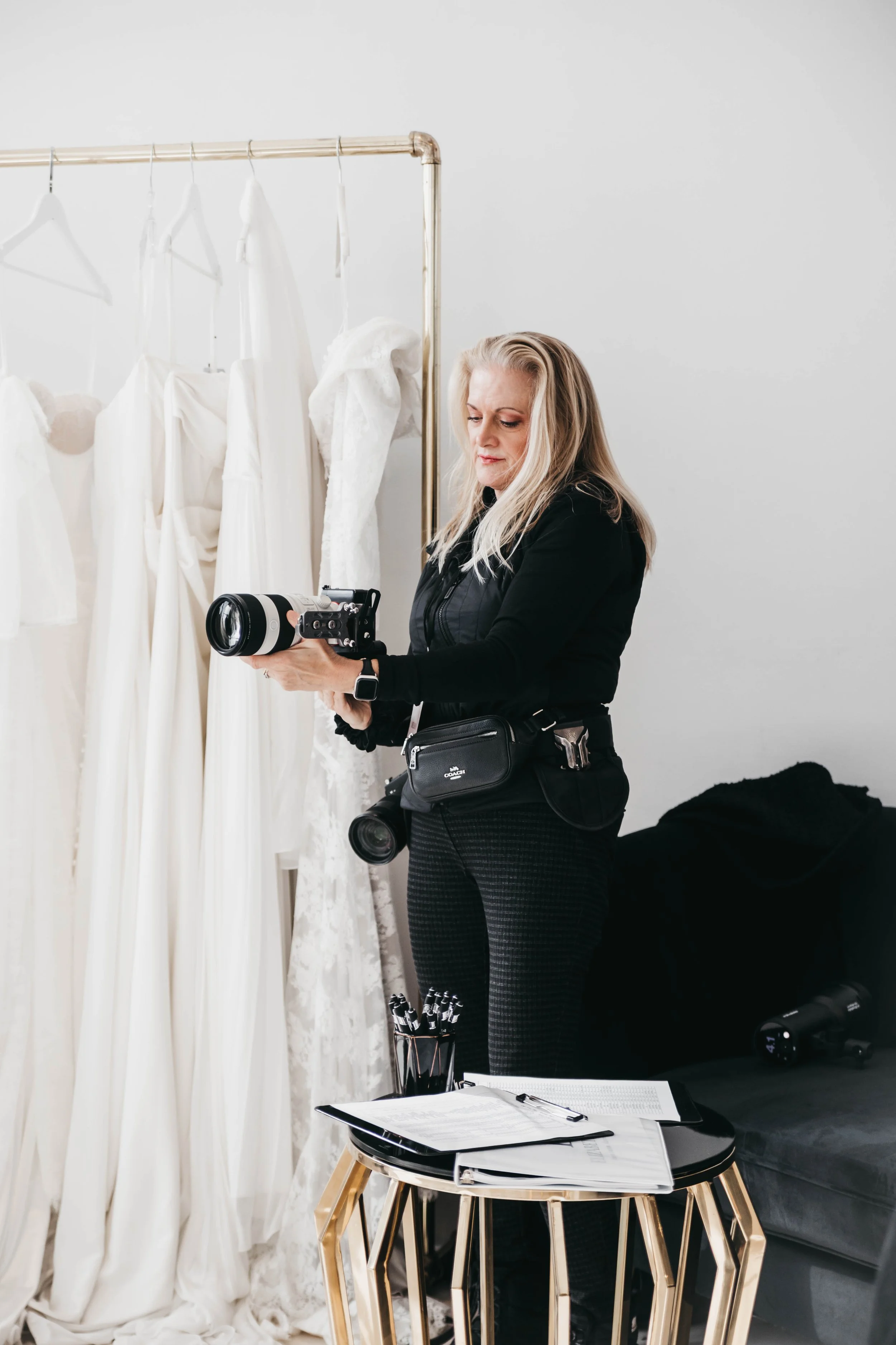 A woman with blonde hair standing indoors, looking at a professional camera in her hands, next to a table with opened notebooks, pens, and clipboards. Behind her is a wardrobe with white dresses hanging.