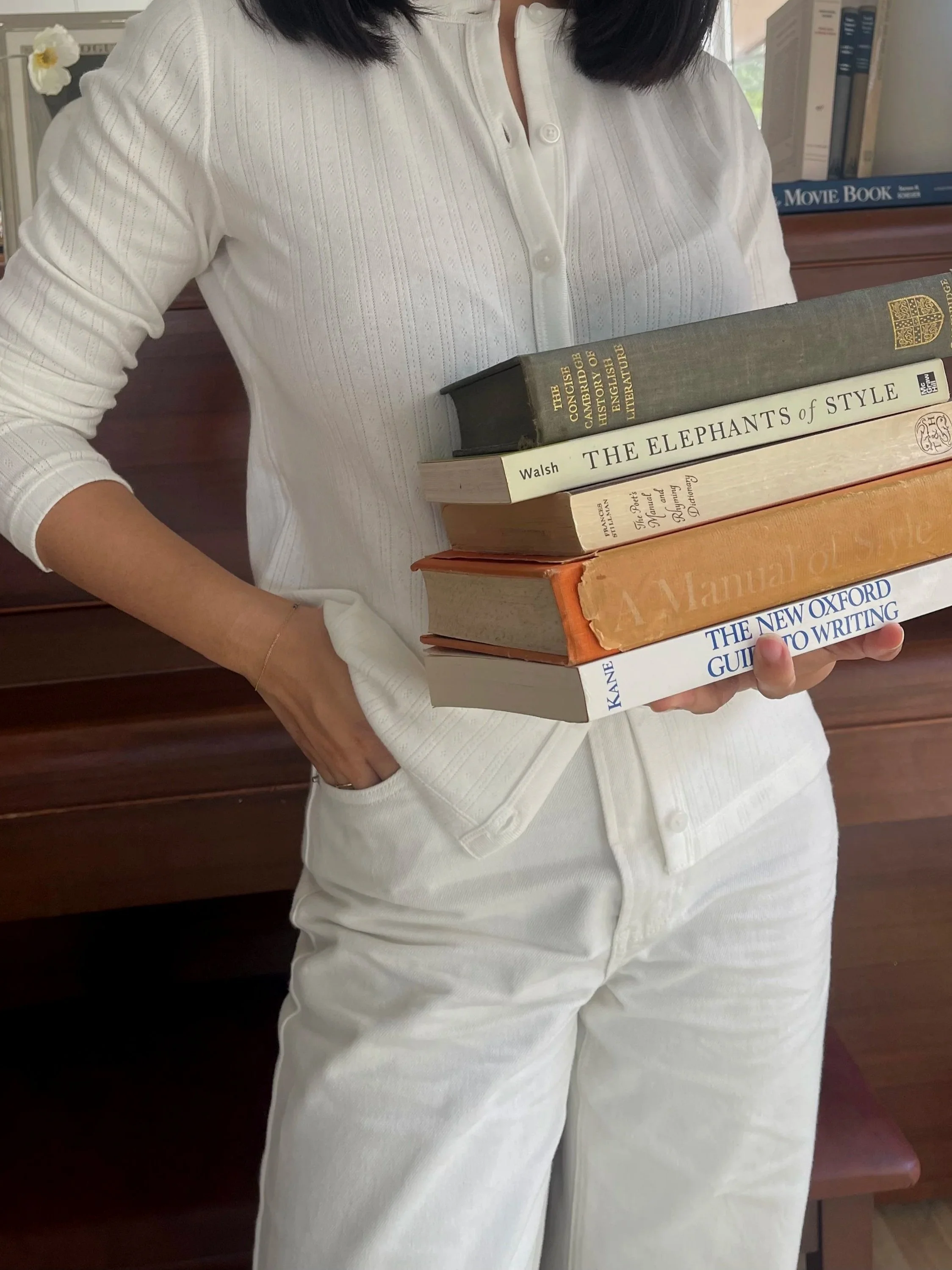 A woman holding a stack of five books in her left hand. She is wearing a white button-up shirt and white pants, and has dark hair. The background includes a wooden surface and bookshelves.