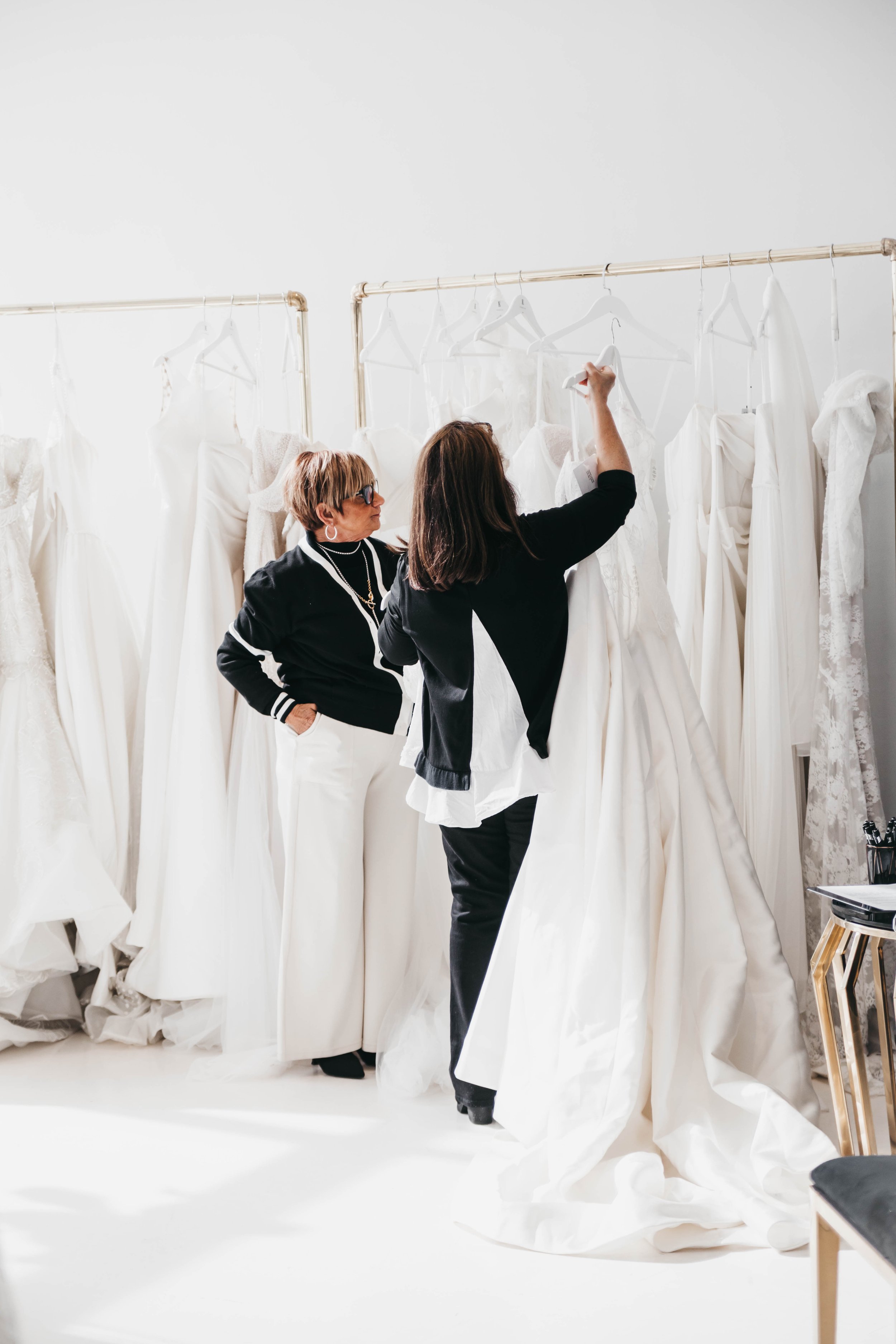 Two women are shopping for wedding dresses in a store. They are looking at white gowns hanging on a rack, with some faced with lace and tulle. The women are engaged in discussion, one holding a hanger.