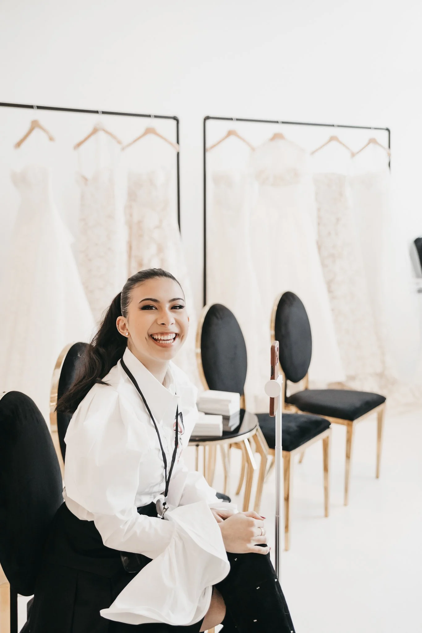 Young woman with dark hair smiling, sitting in a salon with wedding dresses hanging on racks behind her and black chairs.