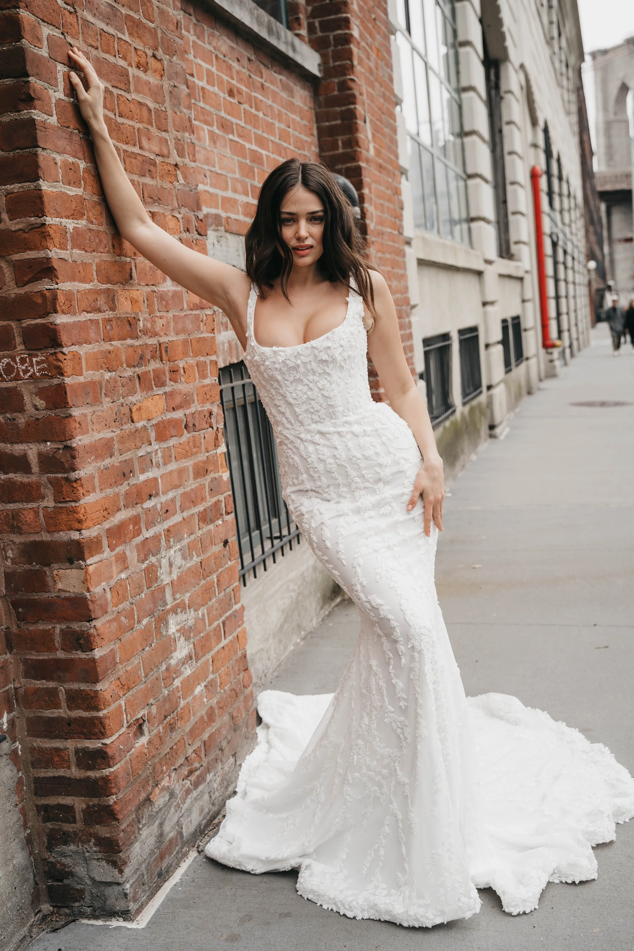 A woman in a white wedding dress leaning against a brick wall on a city sidewalk.
