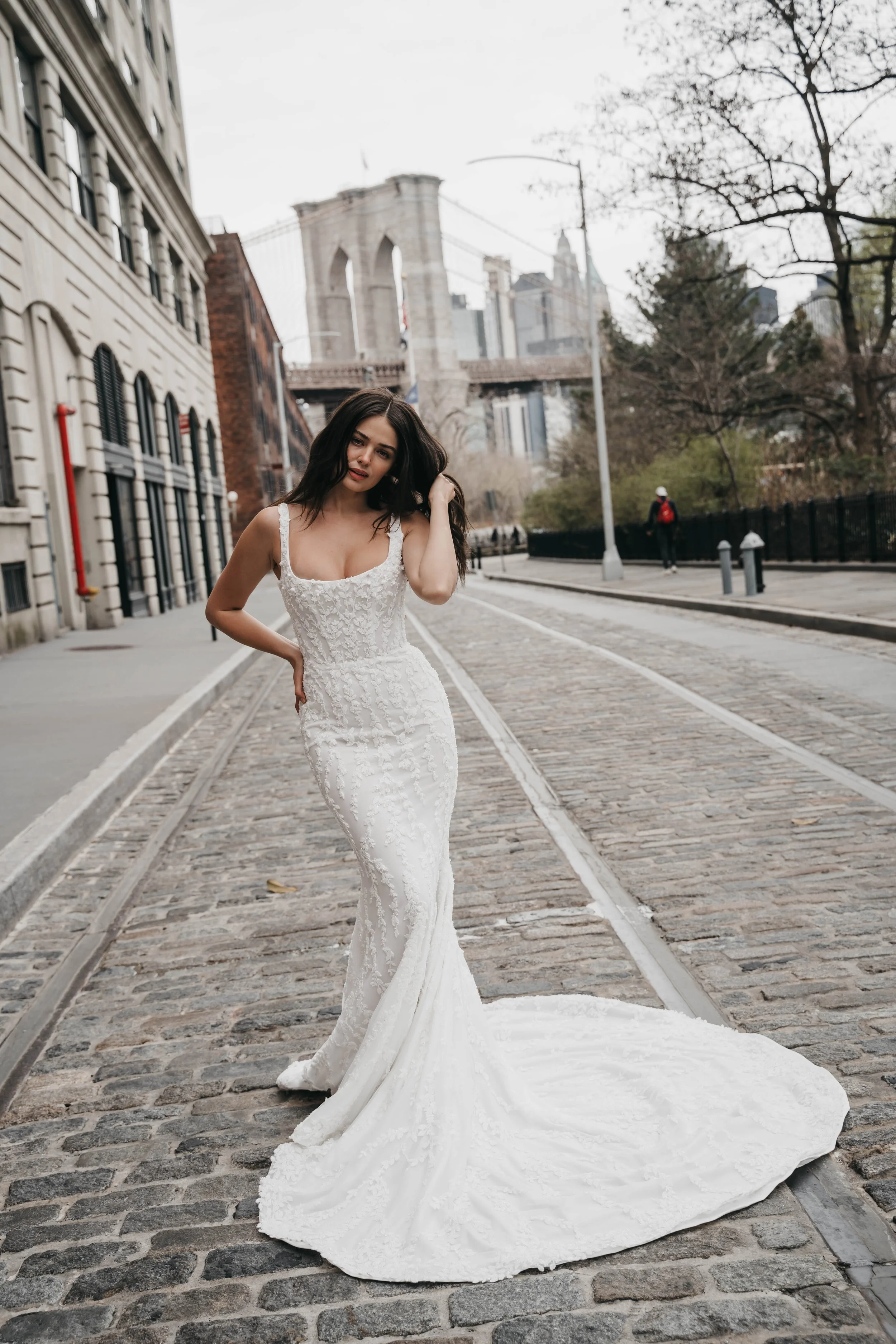 A woman in a white wedding dress standing on cobblestone street near the Brooklyn Bridge in New York City, with bare trees and buildings in the background.