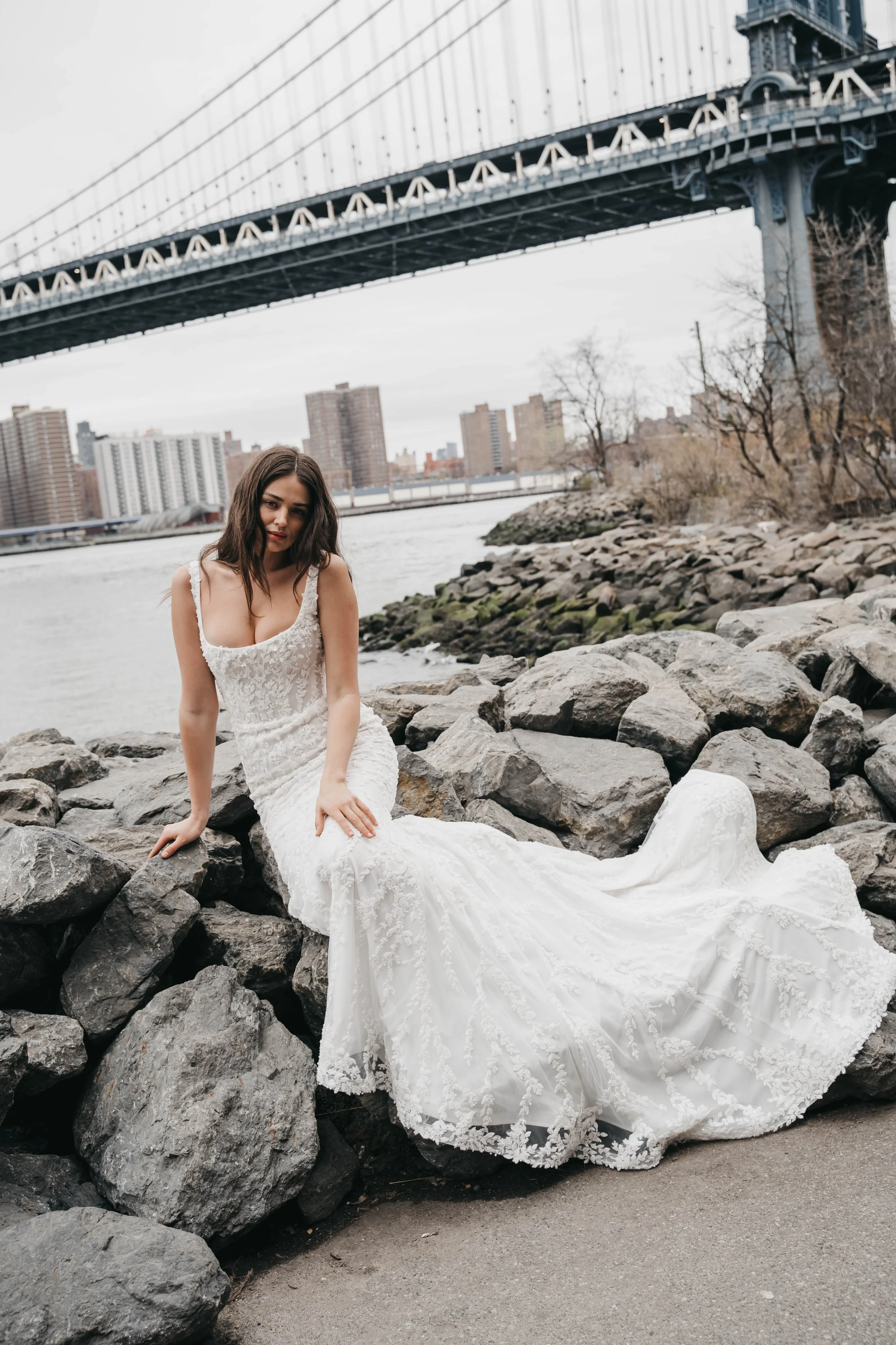 A young woman in a white lace wedding dress sitting on rocks by a river, with city buildings and a bridge in the background.