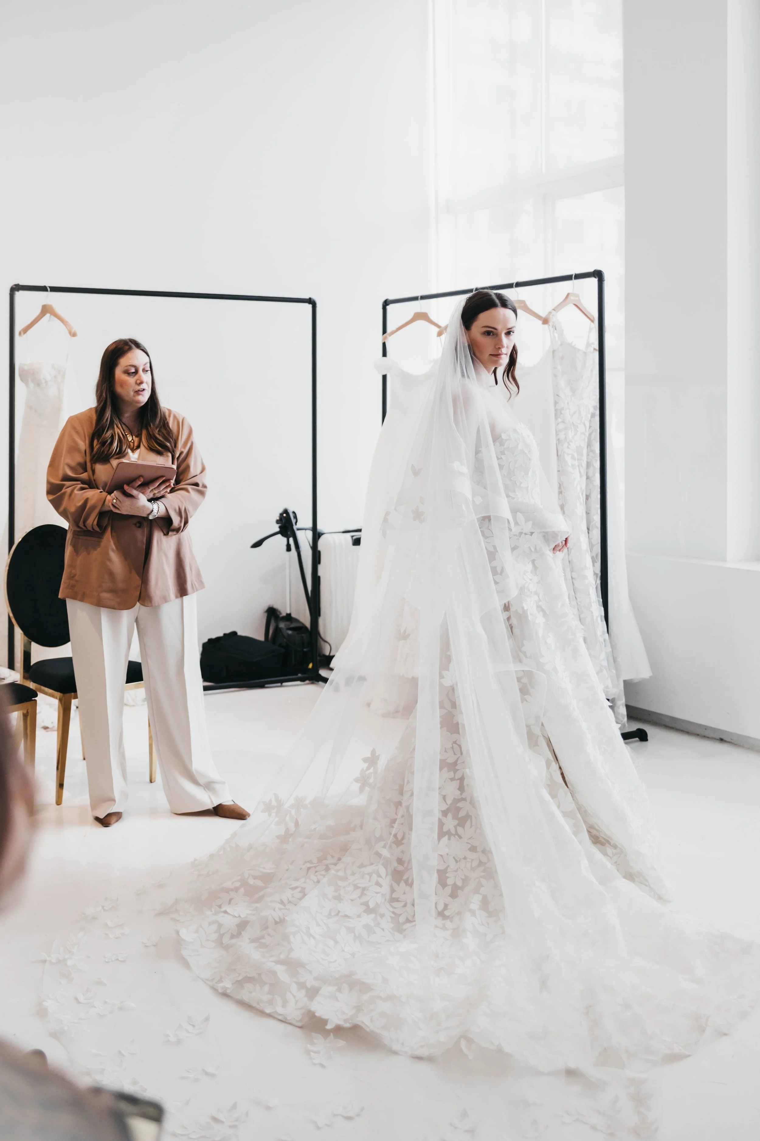 A woman in a wedding dress with a long train and veil looks over her shoulder, standing in front of clothing racks with white dresses. Another woman in beige blazer and white pants holds a tablet and looks towards her, in a bright, white room.