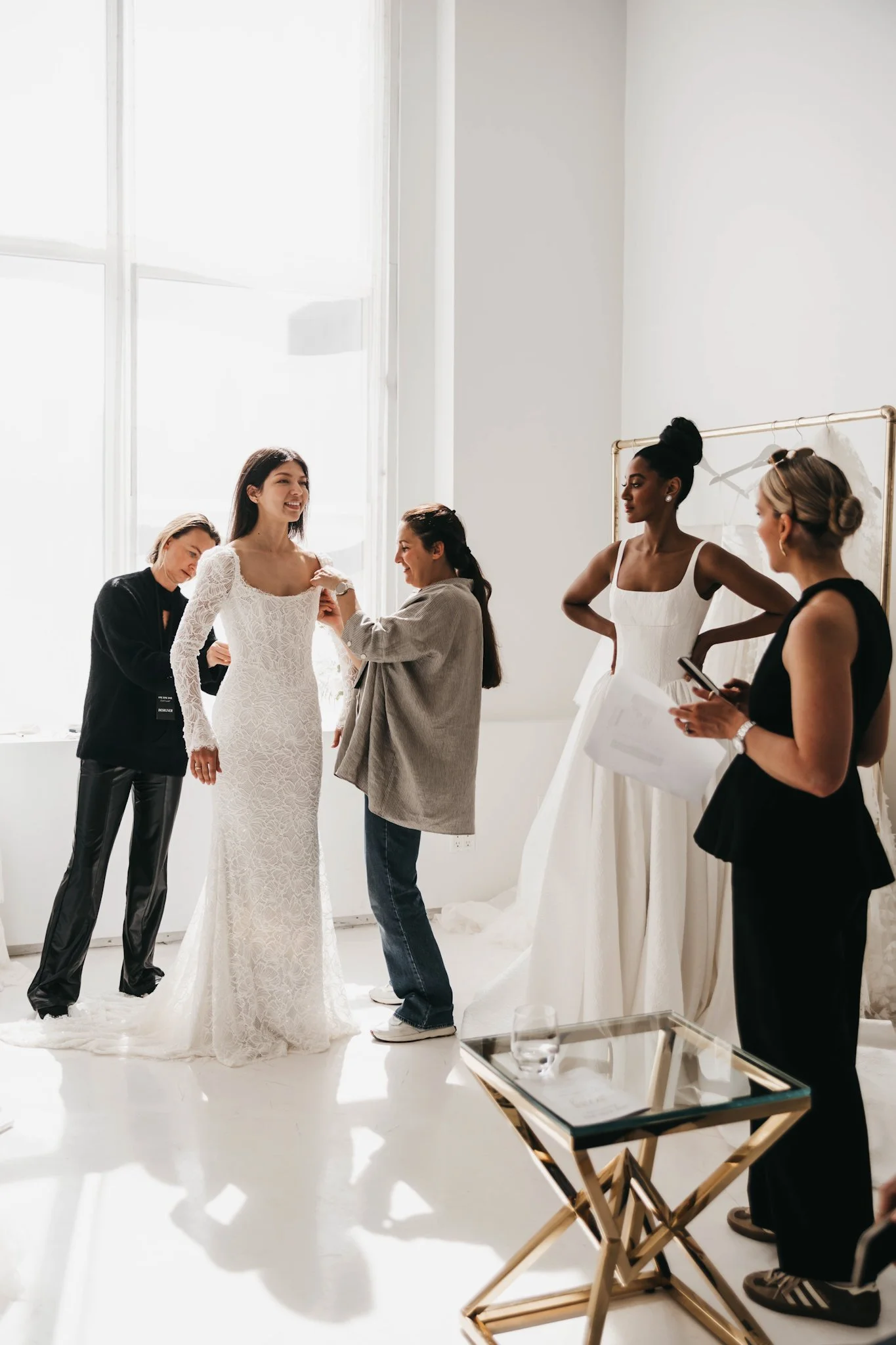 A bride in a white lace wedding gown is having her dress adjusted by a woman in casual attire, while other women, one in a white dress and others in black dresses, look on in a bright, white room with large windows, as part of a wedding dress fitting