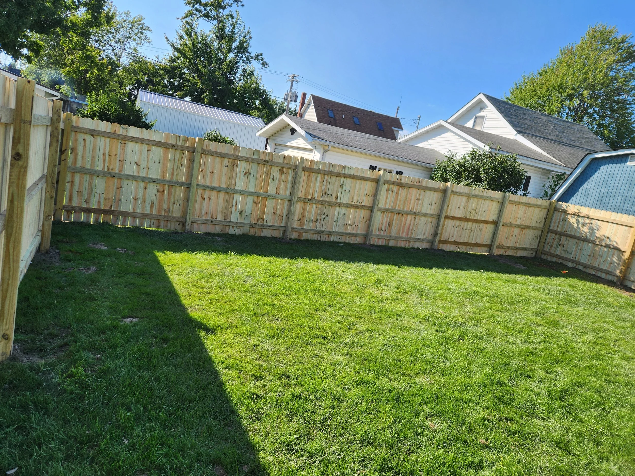 A backyard with a grassy lawn, a tall wooden fence, and several houses with rooftops and trees in the background under a clear blue sky.