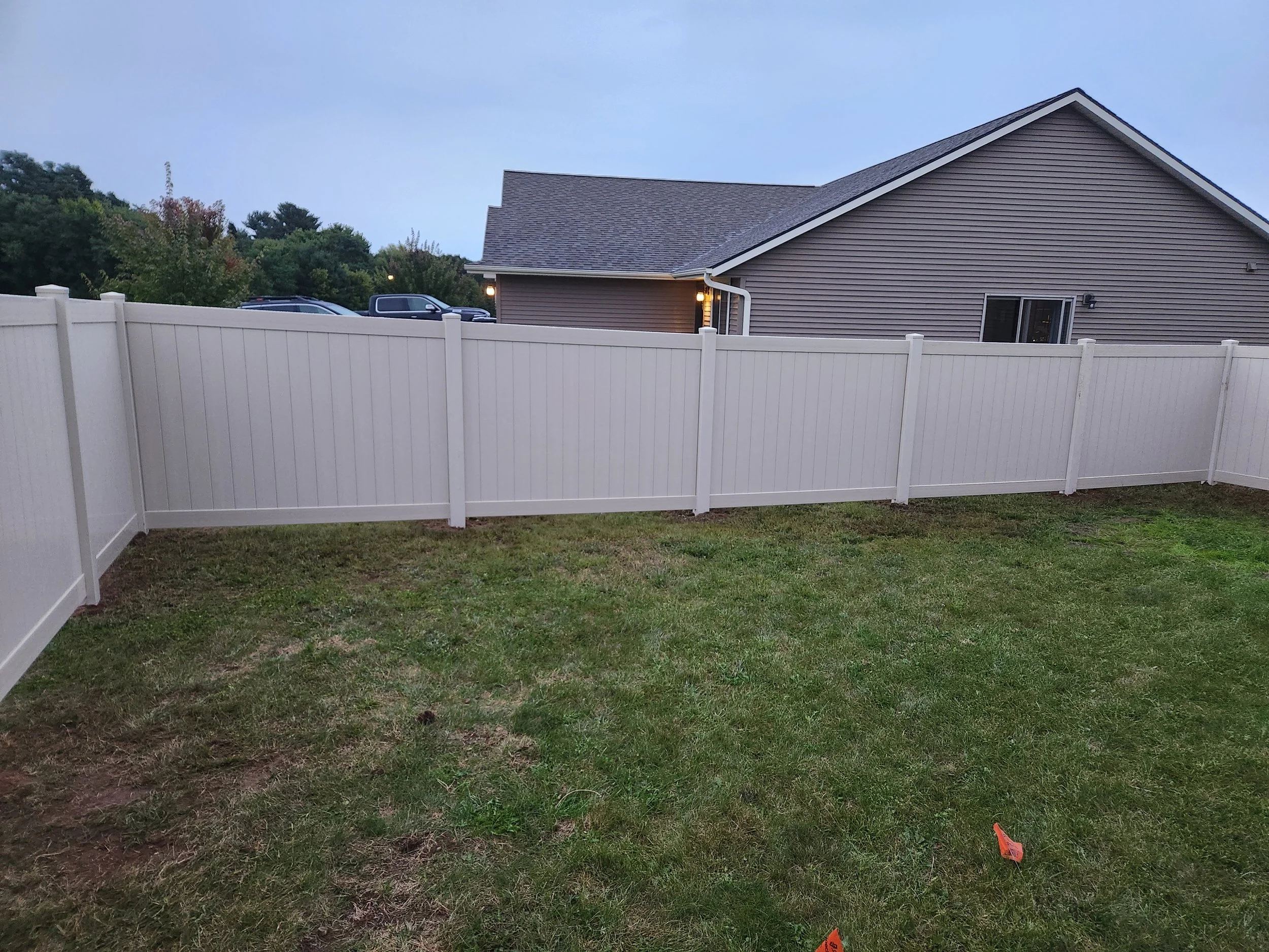 A backyard with green grass and a white privacy fence. In the background, a house with tan siding and a gray roof, with trees and a driveway with parked vehicles. Some yard markers are visible on the grass.