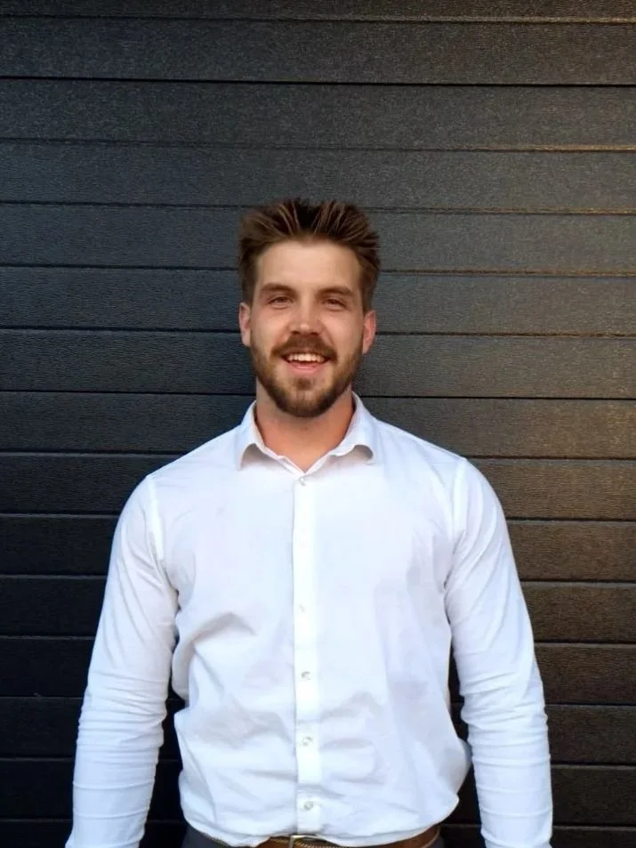 Young man with short brown hair and beard, wearing a white dress shirt, smiling, standing against a dark wooden wall.