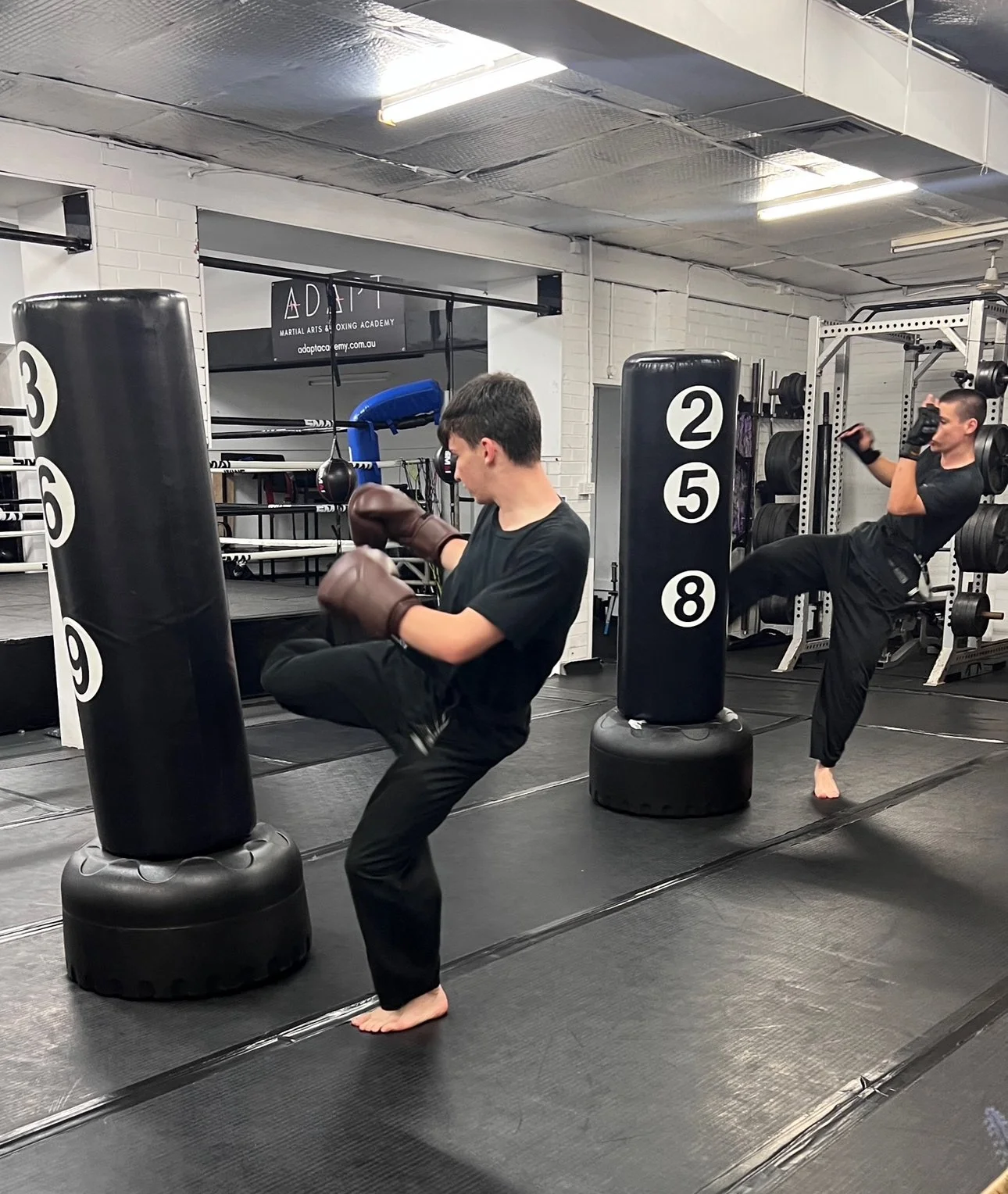 Two individuals practicing martial arts in a gym, one with brown gloves kicking a standing punching bag and the other doing a high kick near weight racks.