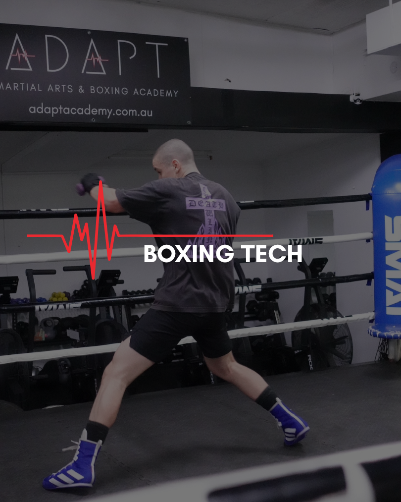 A person boxing in a gym, standing in a boxing stance inside a ring with punching gloves on, surrounded by gym equipment, with a sign that says "ADAPT Martial Arts & Boxing Academy" in the background.