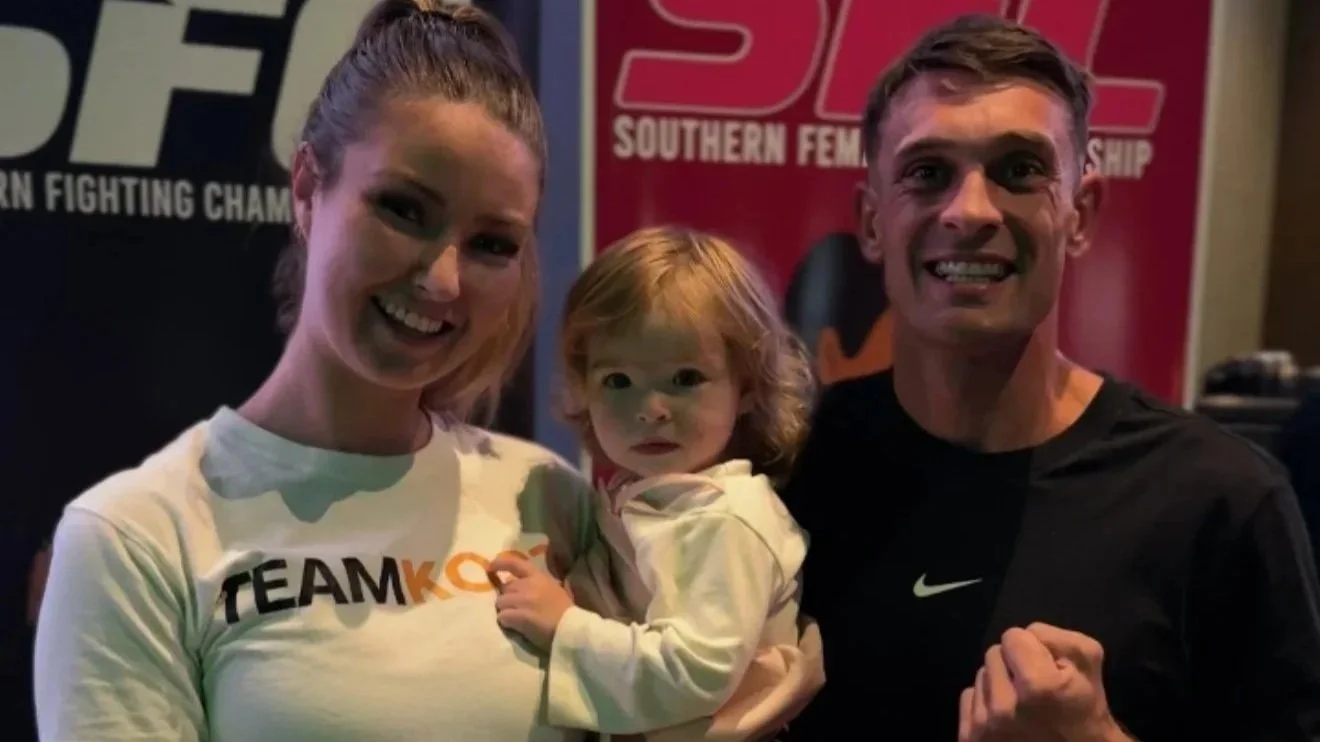 A woman, a young girl, and a man smiling and posing together at an event with banners in the background, one reading 'SOUTHERN FIGHTING' and the other 'TEAM KOR' on the woman's shirt.