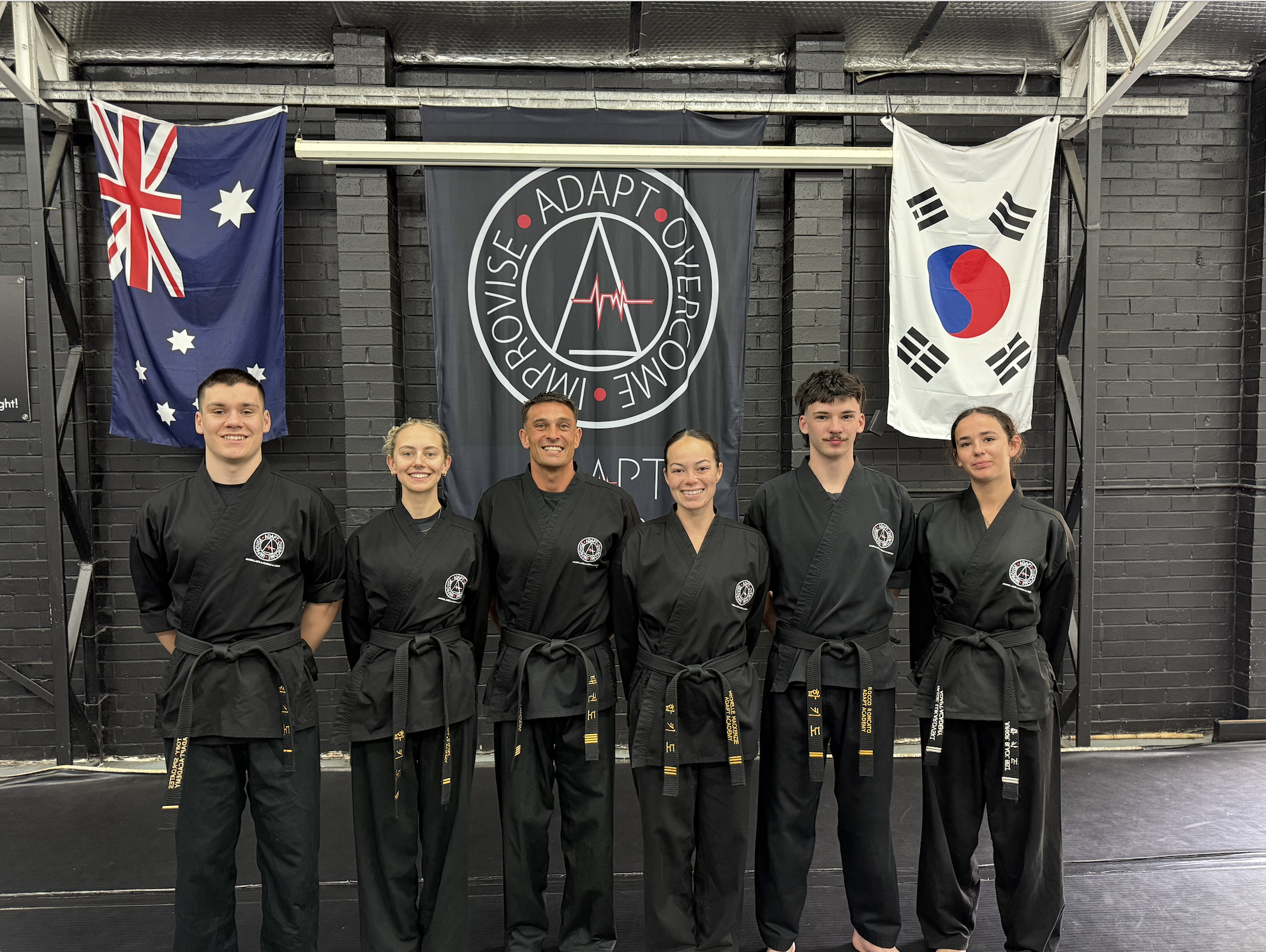Group of six martial artists in black uniforms standing in a dojo with flags from Australia, South Korea, and a circular banner with training logo behind them.