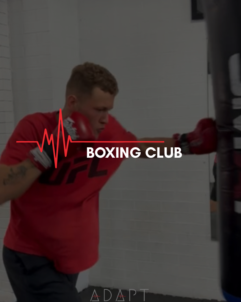 A young man in a red t-shirt and boxing gloves practicing strikes on a punching bag in a boxing gym.