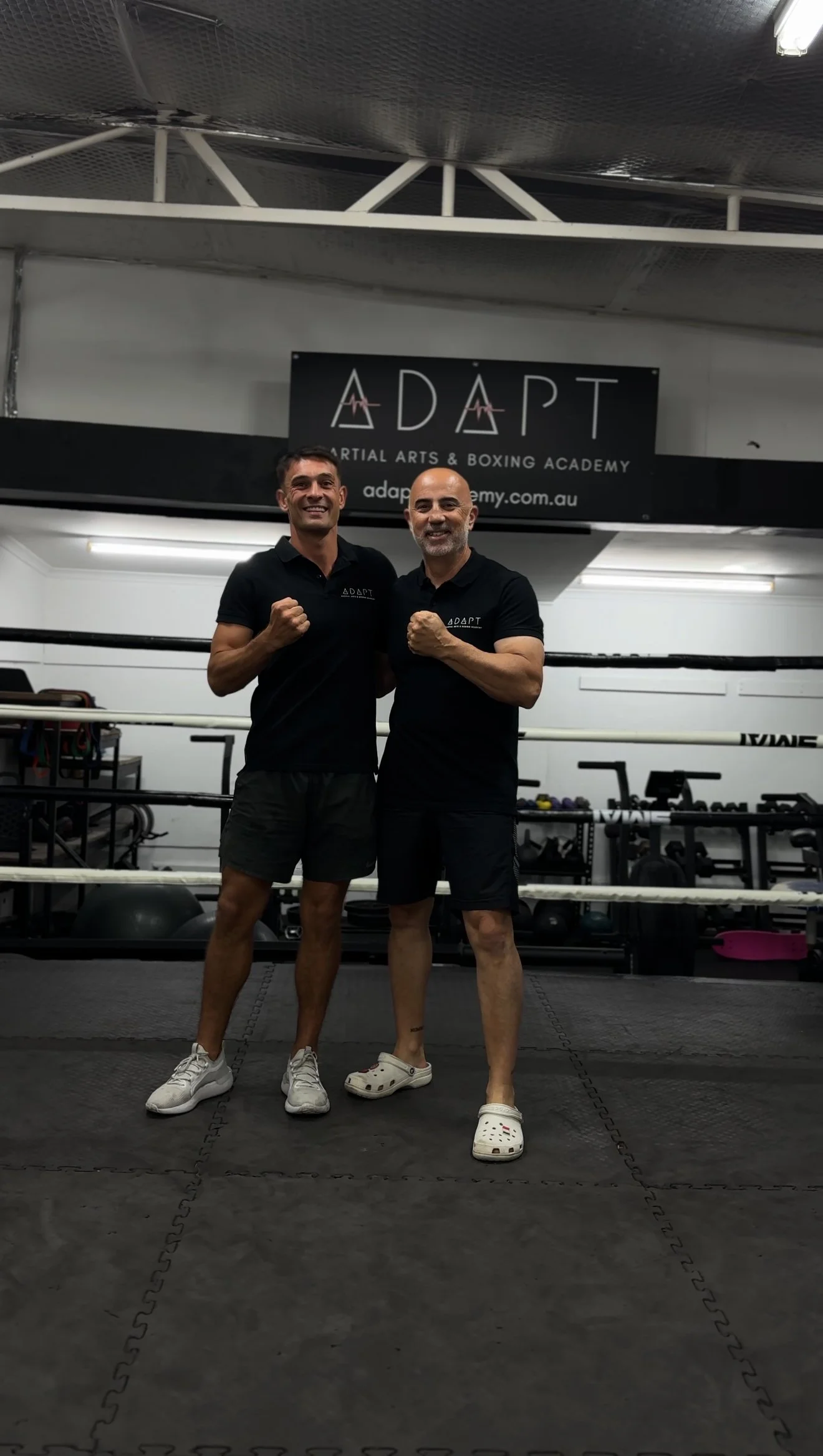 Two men standing inside a martial arts and boxing gym, smiling and flexing their arms. They are both wearing black polo shirts with the gym's logo, 'ADAPT,' and black shorts. Behind them is a boxing ring and gym equipment. A sign with the gym's name 'ADAPT Martial Arts & Boxing Academy' is visible on the wall.