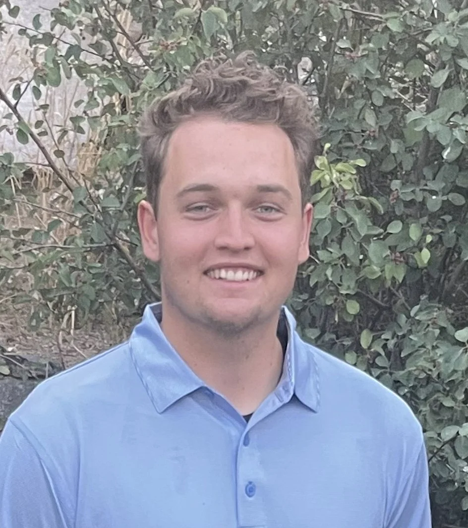 A young man with short curly hair smiling and wearing a light blue collared shirt, standing outdoors in front of bushes with green leaves.