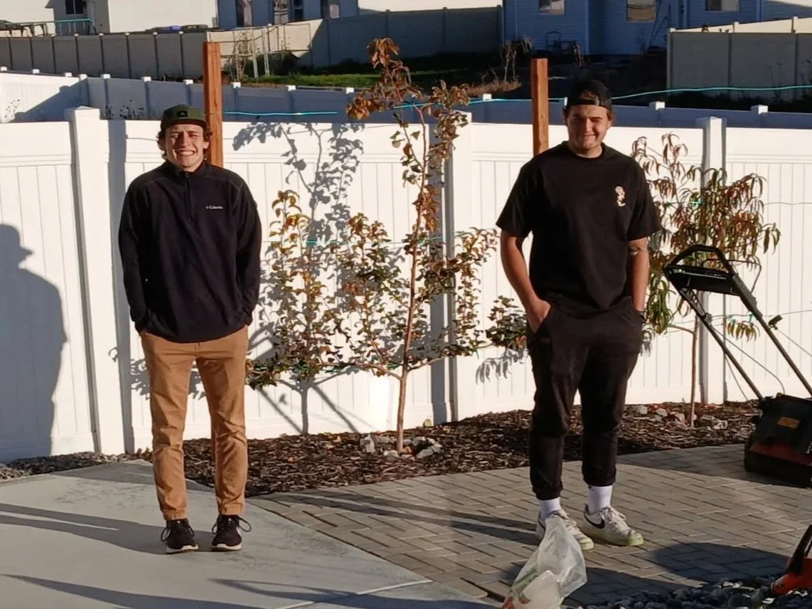 Two young men standing outside near a white fence, smiling with trees and a garden in the background.