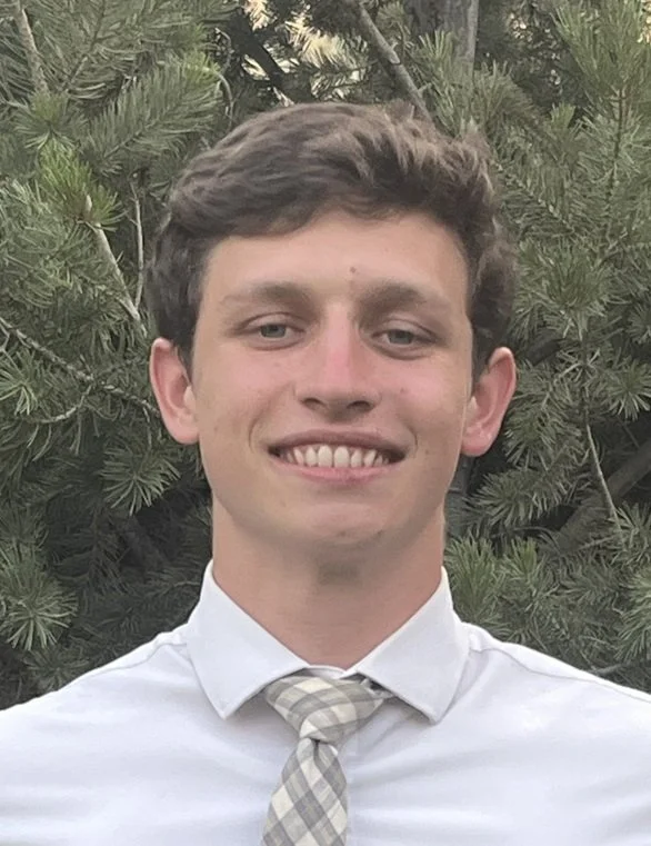Young man with short brown hair and light skin, smiling, wearing a white dress shirt and a light-colored striped tie, standing outdoors with green pine tree branches in the background.