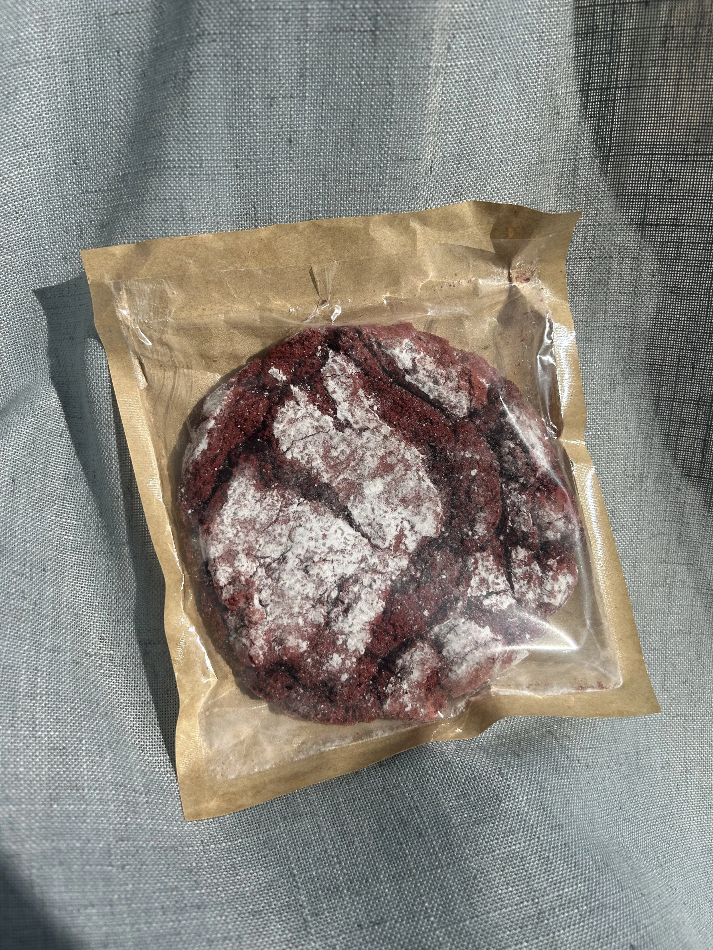 Photo of a big red velvet packaged in a partial brown bag with a clear plastic window that can clearly see the entire cookie
