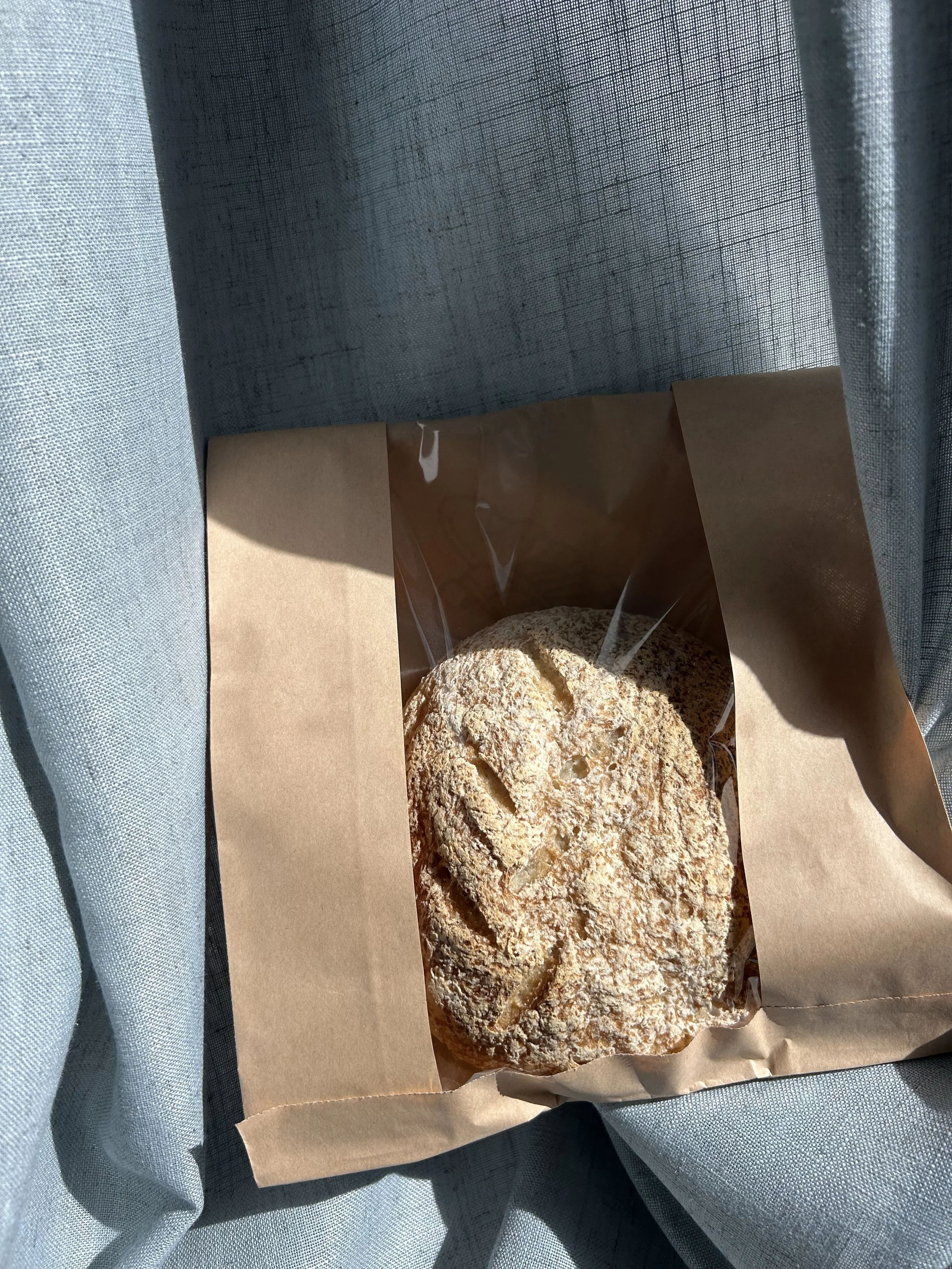 Round bread loaf packaged in a brown bag with a clear window, sitting in the sunlight