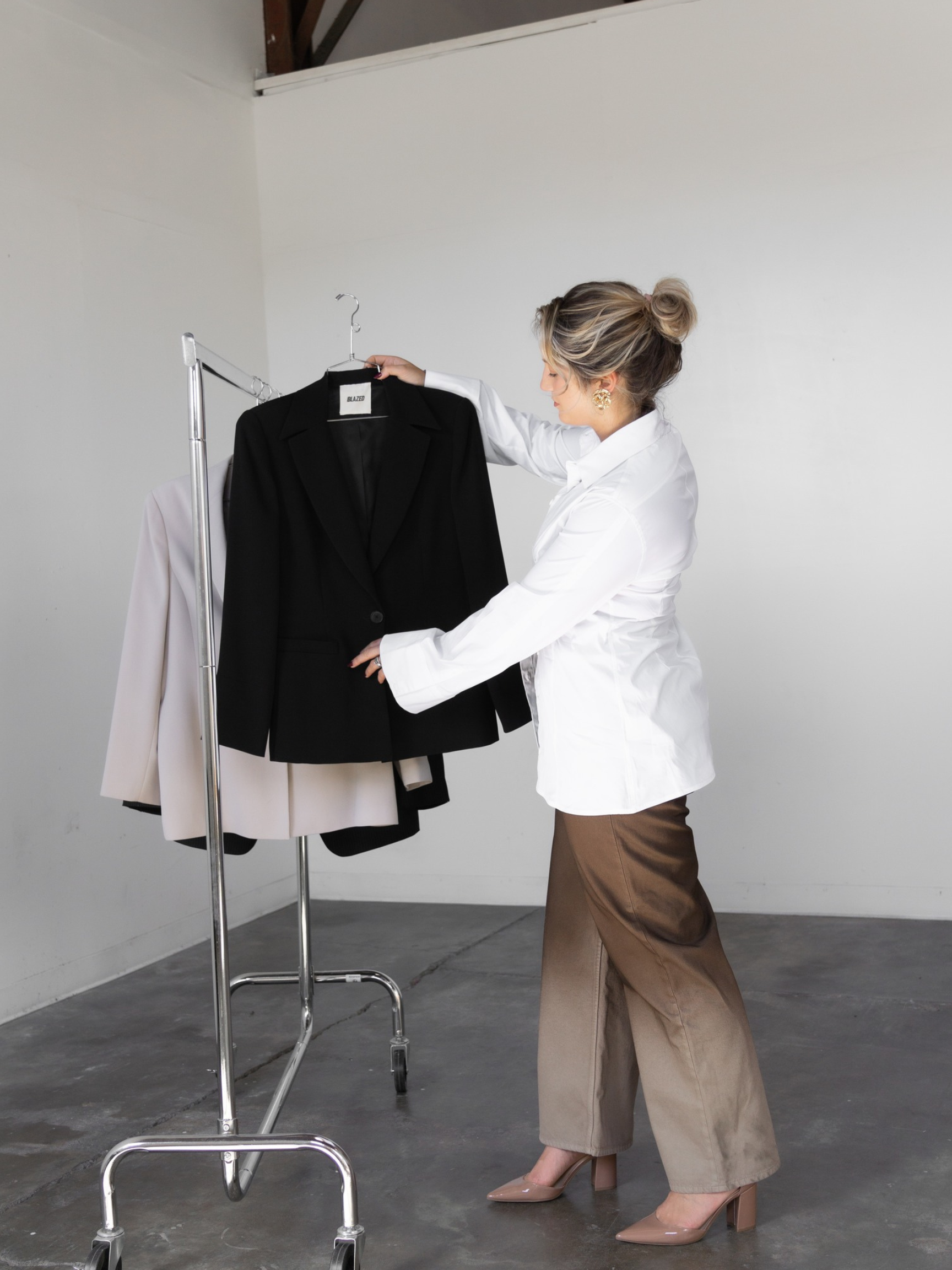 A woman wearing a white blouse, beige wide-leg pants, and nude high heels is looking at black, white, and beige blazers hanging on a metal clothing rack in front of a plain white wall.