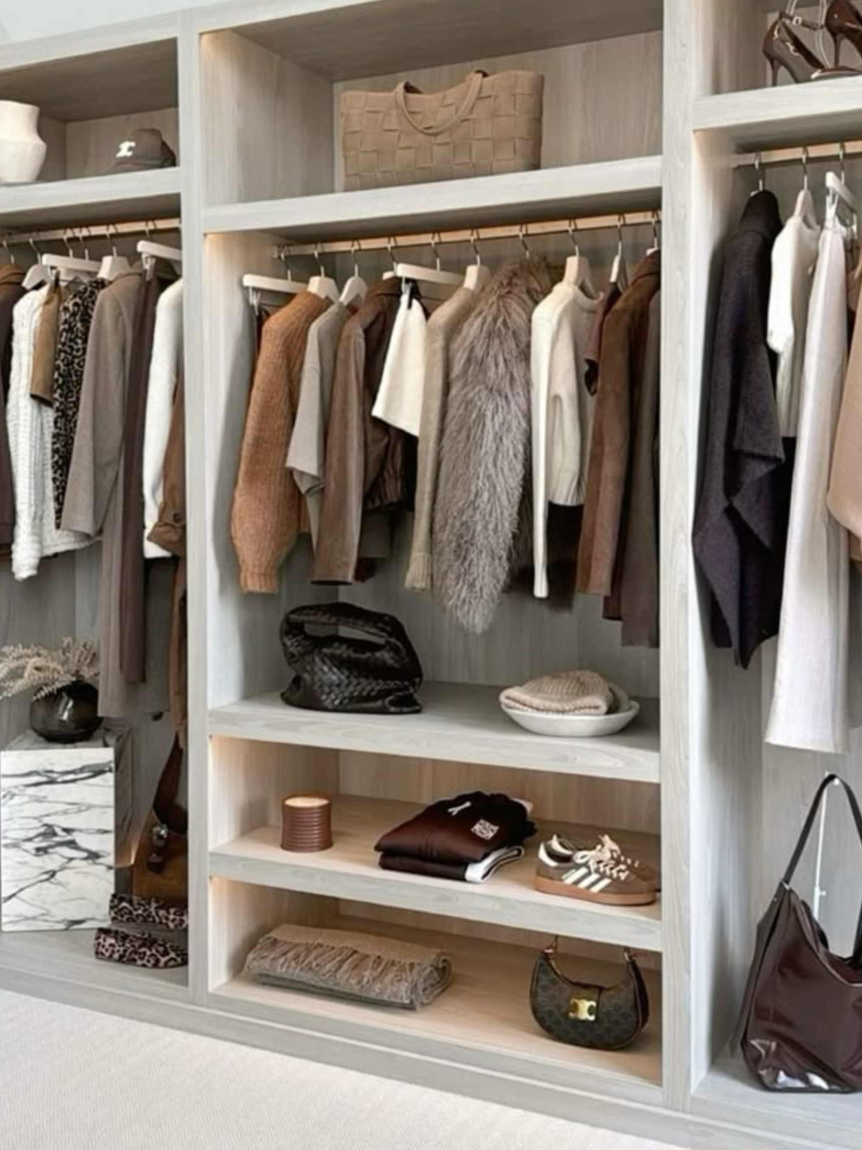 A closet with open shelving and hanging clothes in neutral and earth tones, including leather and faux fur pieces, with handbags, shoes, and folded clothing stored on the shelves.