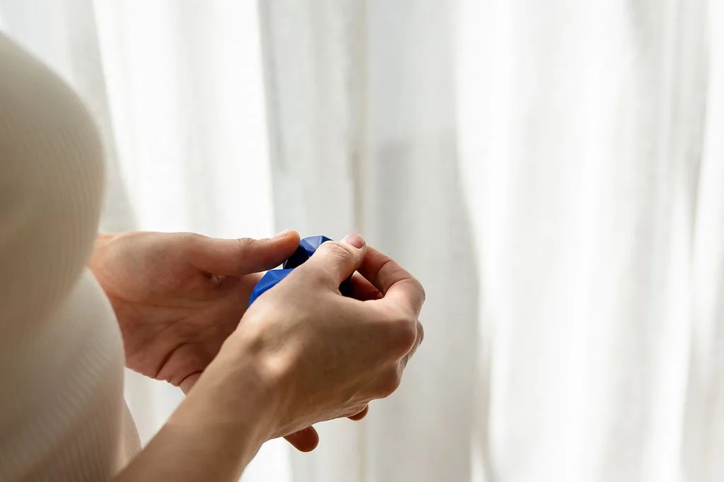 Close-up of a person's hands holding a blue remote control, with a blurred white curtain in the background.