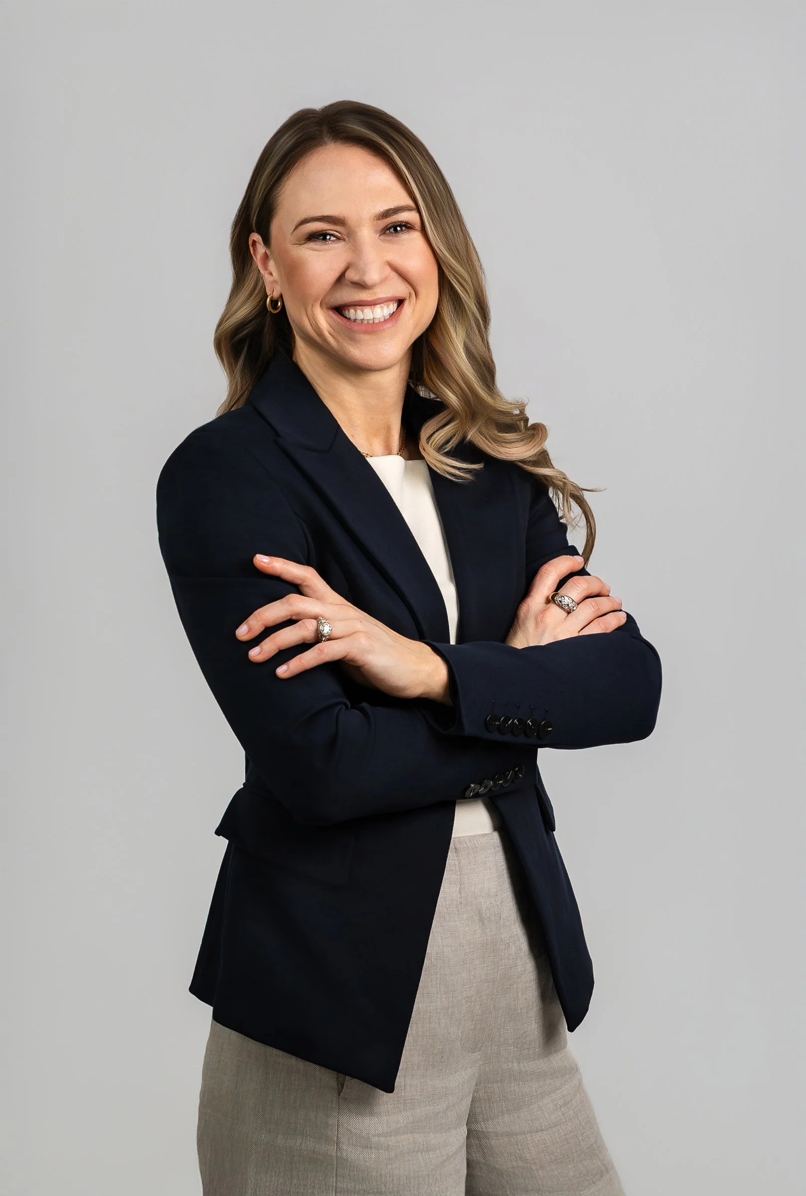 A woman with light brown, wavy hair smiling confidently, wearing a dark blazer over a white top, with crossed arms, standing against a plain light gray background.