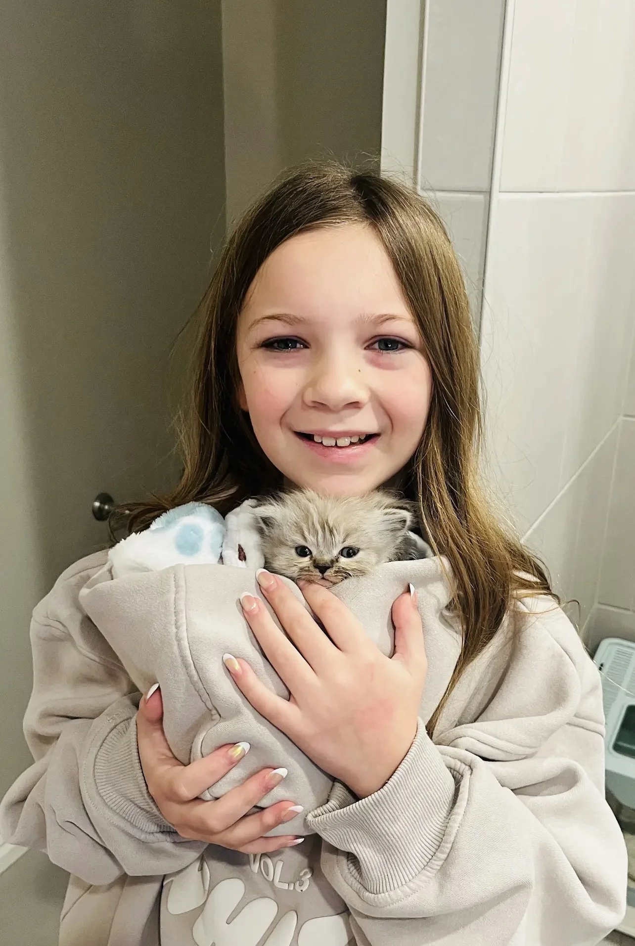 A young girl with long brown hair and a beige sweatshirt is smiling and holding a small gray kitten in a soft beige blanket, in an indoor setting.