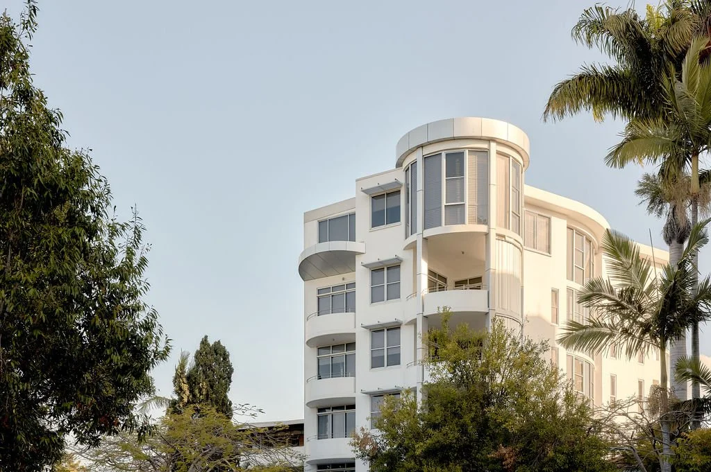 CPD. A white modern multi-story apartment building with curved balconies, surrounded by trees and palm trees under a clear sky.