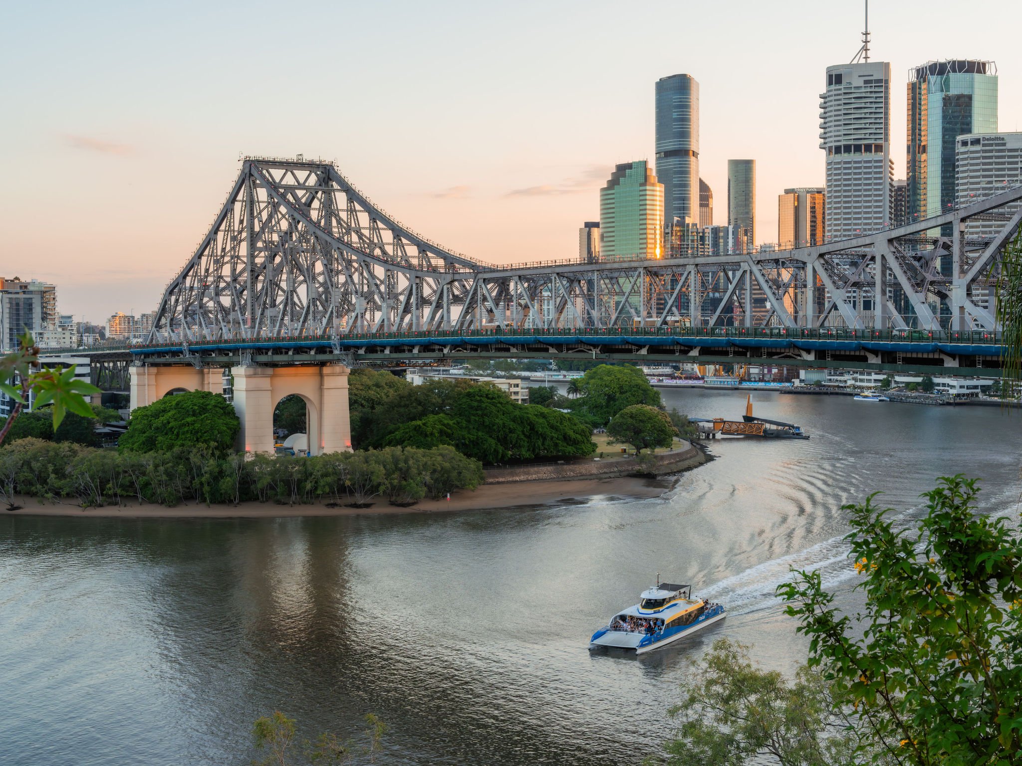 A city skyline with tall modern buildings and skyscrapers, a bridge crossing over a river, and a boat sailing on the water during sunset.