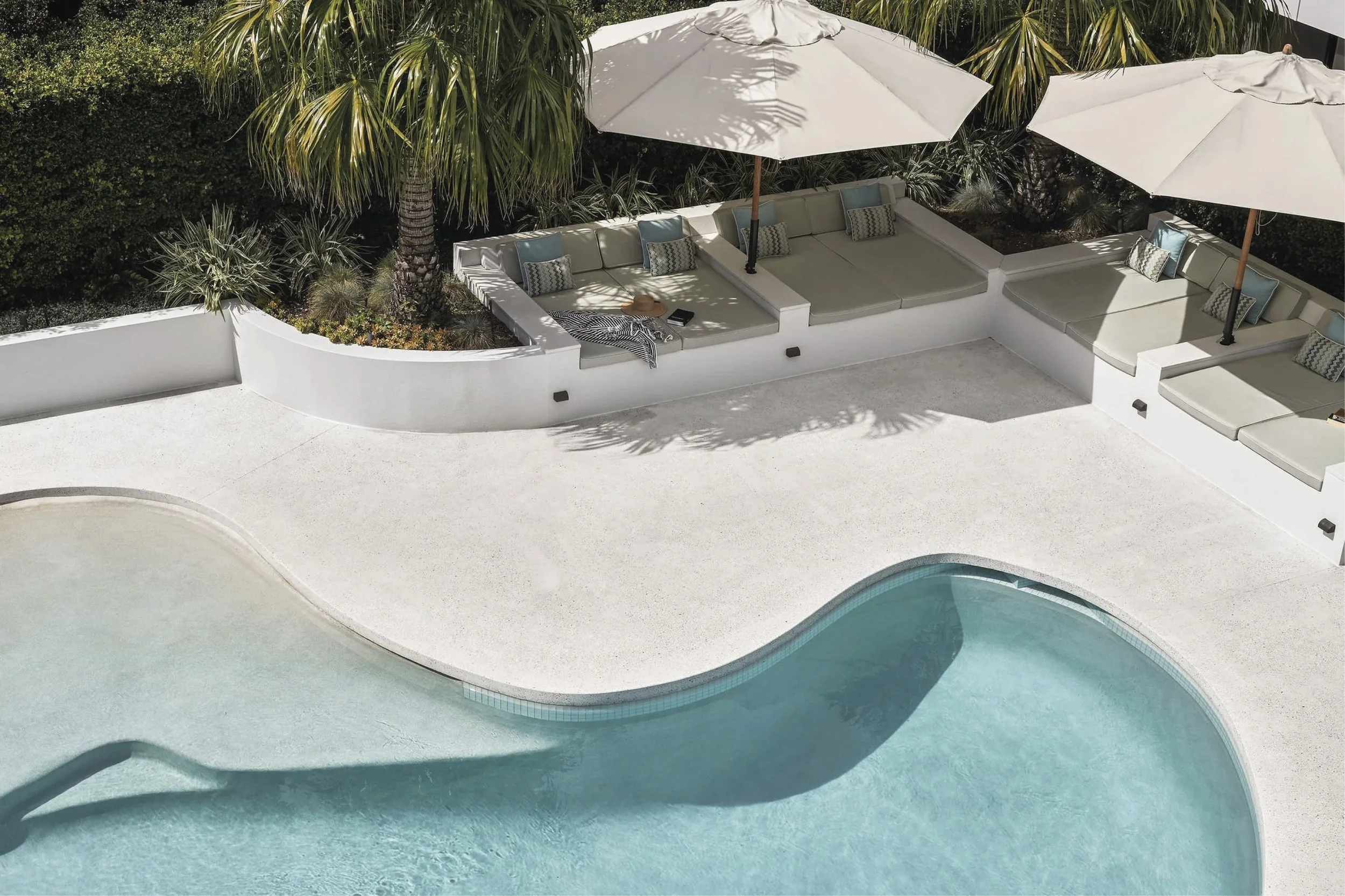 Favell - Empty swimming pool and outdoor seating area with umbrellas, cushions, and palm trees.