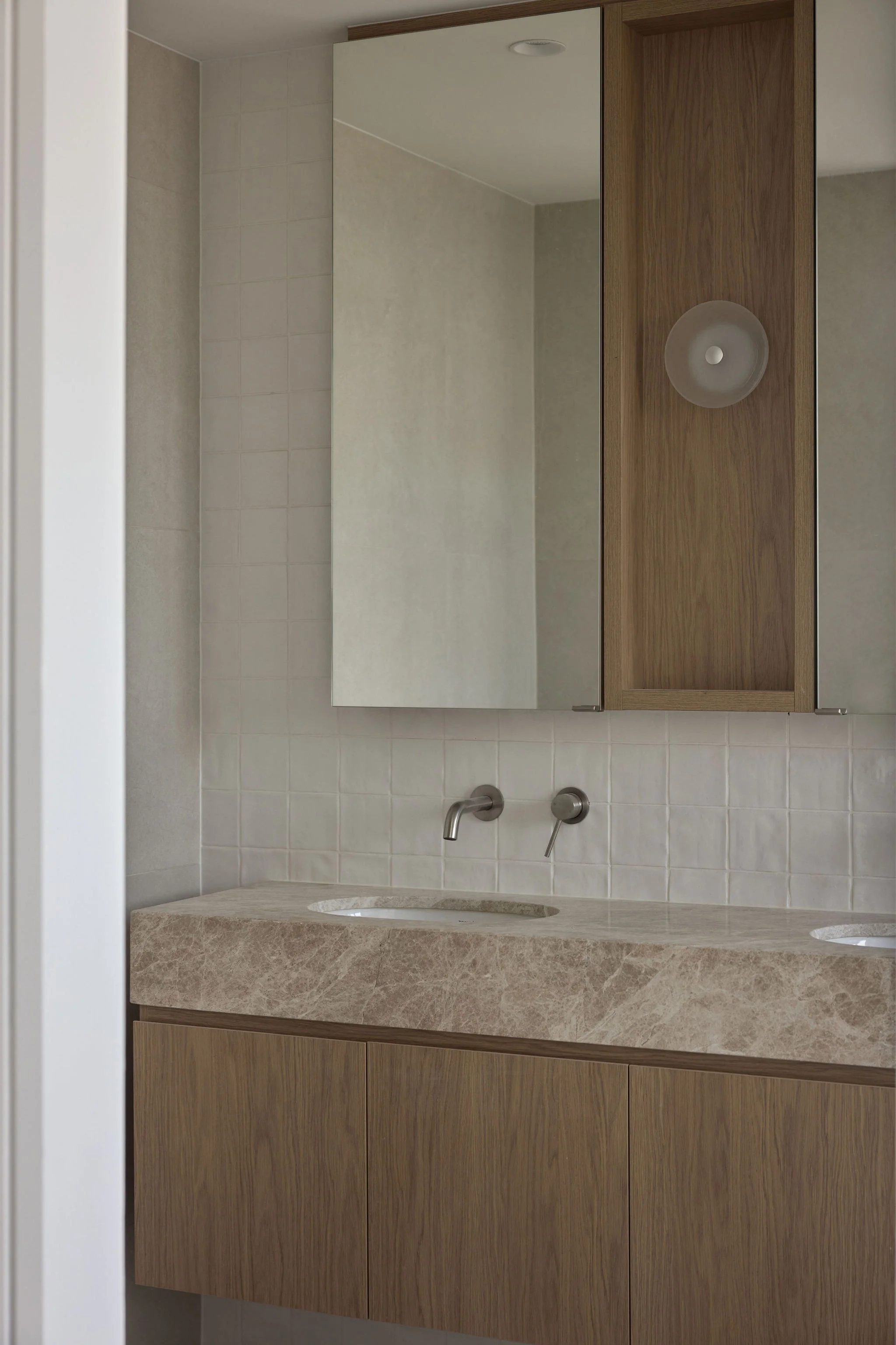 LEDA .A bathroom vanity with a marble countertop, a large mirror, and wooden cabinetry. There are two wall-mounted faucets above the sinks, and a wooden cabinet with a frosted glass door is mounted above the countertop. 793 Brunswick St, New Farm