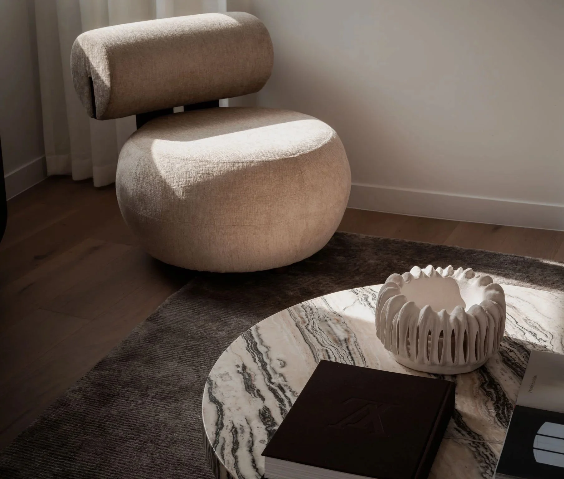 A beige, modern, rounded chair with a curved back rest in a minimalistic interior setting. The chair is next to a window with curtains, and there is a marble coffee table with a decorative white sculpture on top. The room has wooden flooring and a neutral color palette.