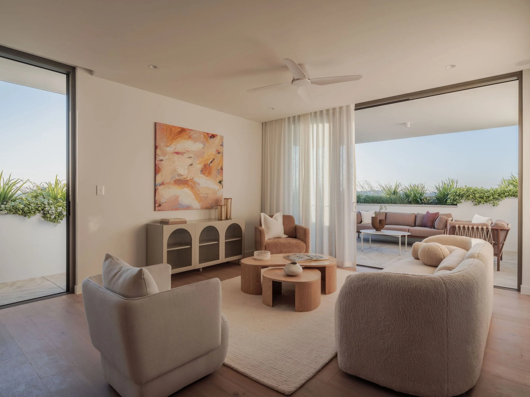 LUMIA Modern living room with large floor-to-ceiling windows, white walls, beige furniture including armchairs and a sofa, wooden coffee tables, an abstract painting on the wall, and curtains. Green plants outside the window.