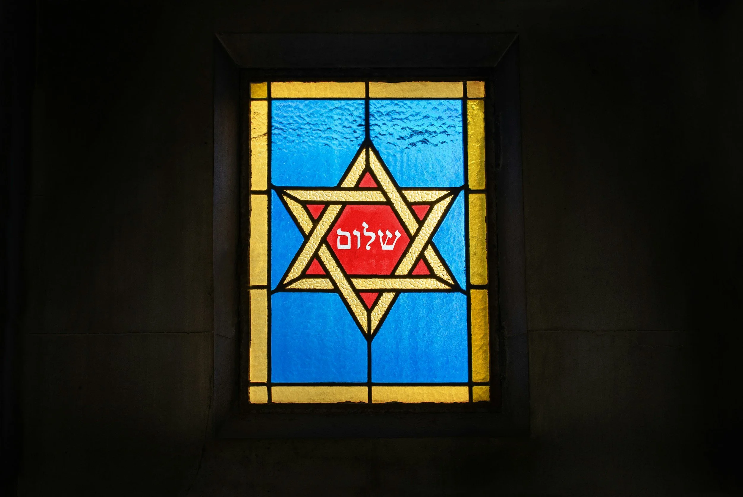 Colorful stained glass window featuring a Star of David with Hebrew word 'Shalom' in the center, surrounded by yellow, blue, red, and black glass.