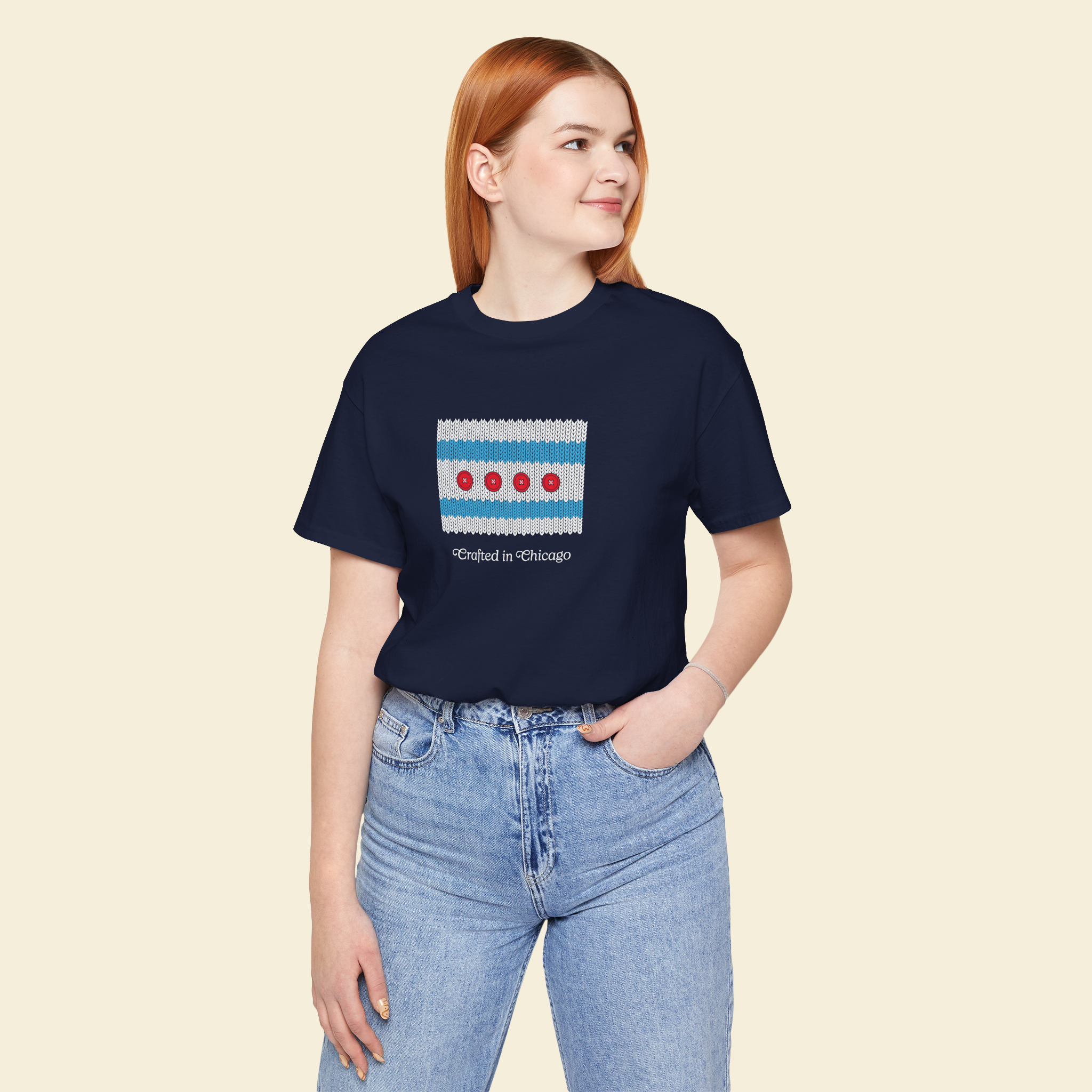 A young woman with red hair wearing a navy t-shirt with a Chicago city flag design and the text "Crafted in Chicago," and light blue jeans, standing against a beige background.