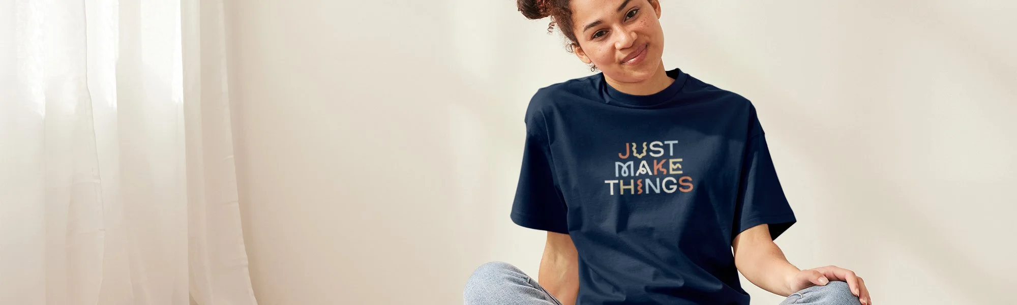 A young mixed race woman with freckles and her curly hair in space buns wears a CrafterCore "Just Make Things" t-shirt while sitting cross-legged in a white studio