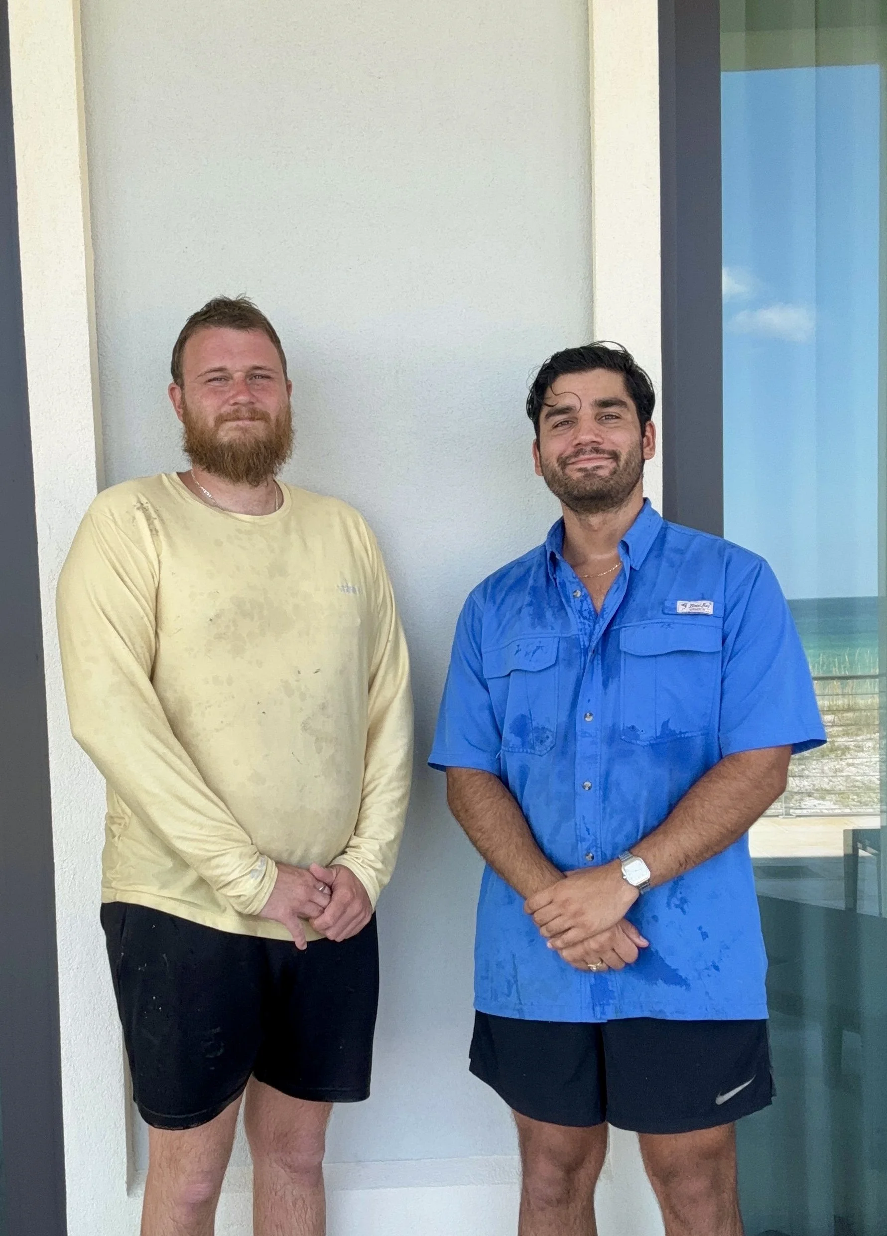 Two men standing outside near a glass window, one wearing a yellow shirt with wet spots and black shorts, the other wearing a blue shirt and black shorts, both smiling.