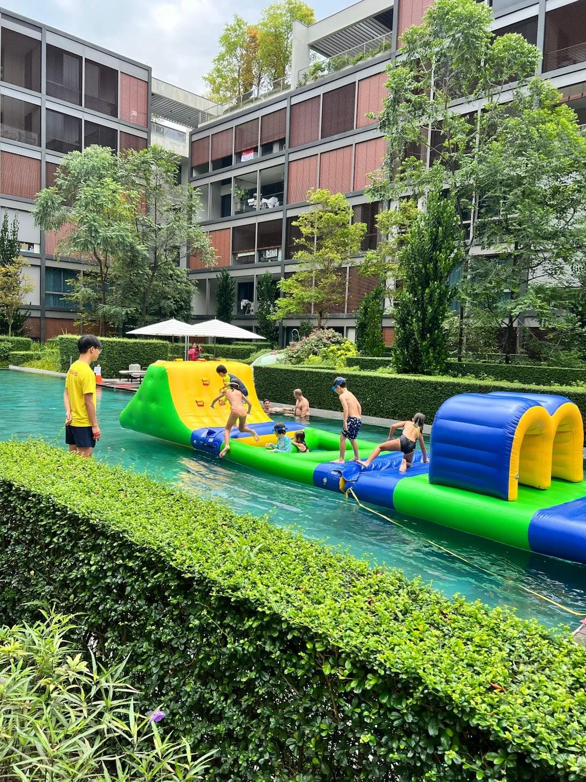 Children and adults playing on a large inflatable water slide in a swimming pool surrounded by greenery and tall apartment buildings.