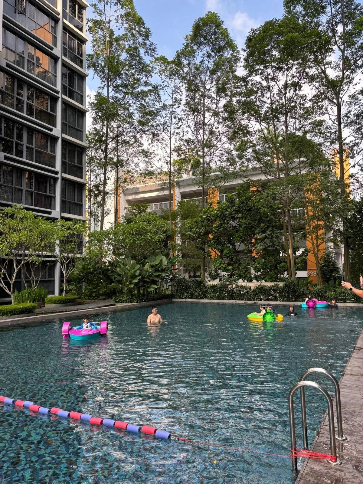 People swimming and floating on inflatable toys in a swimming pool surrounded by tall trees and modern buildings.