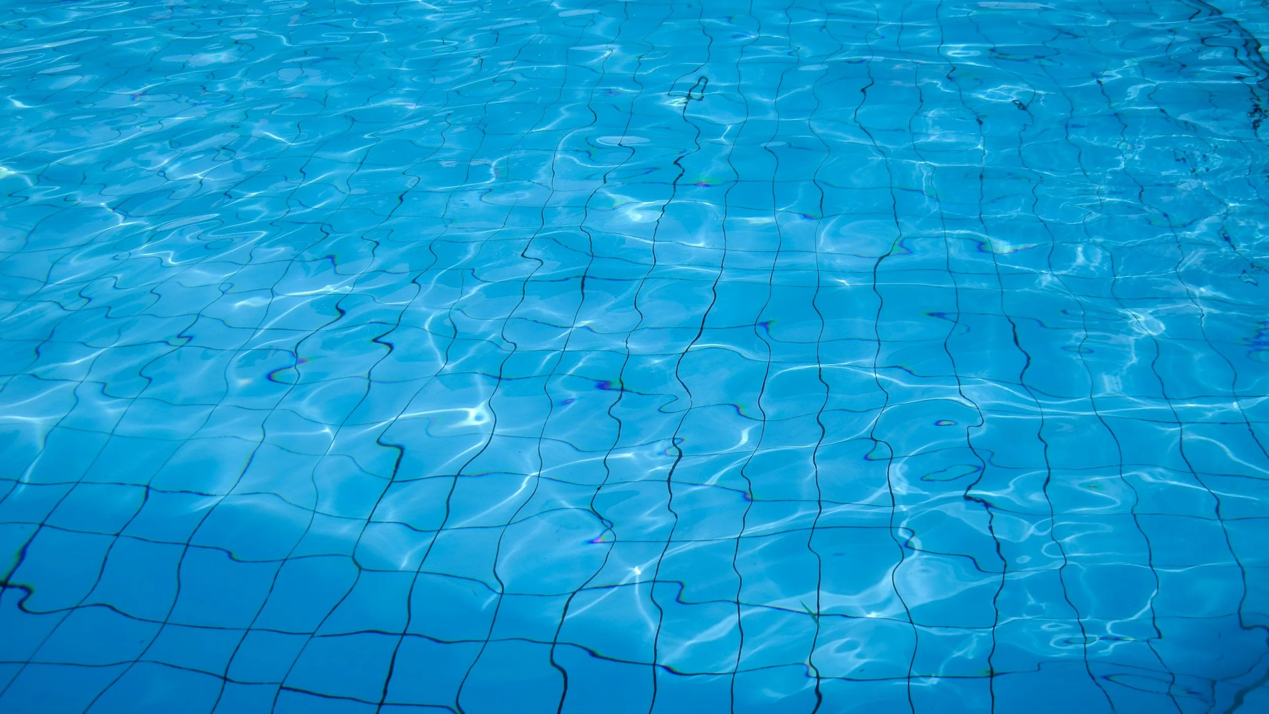 Photo of a swimming pool with blue water and dark lane lines on the bottom.