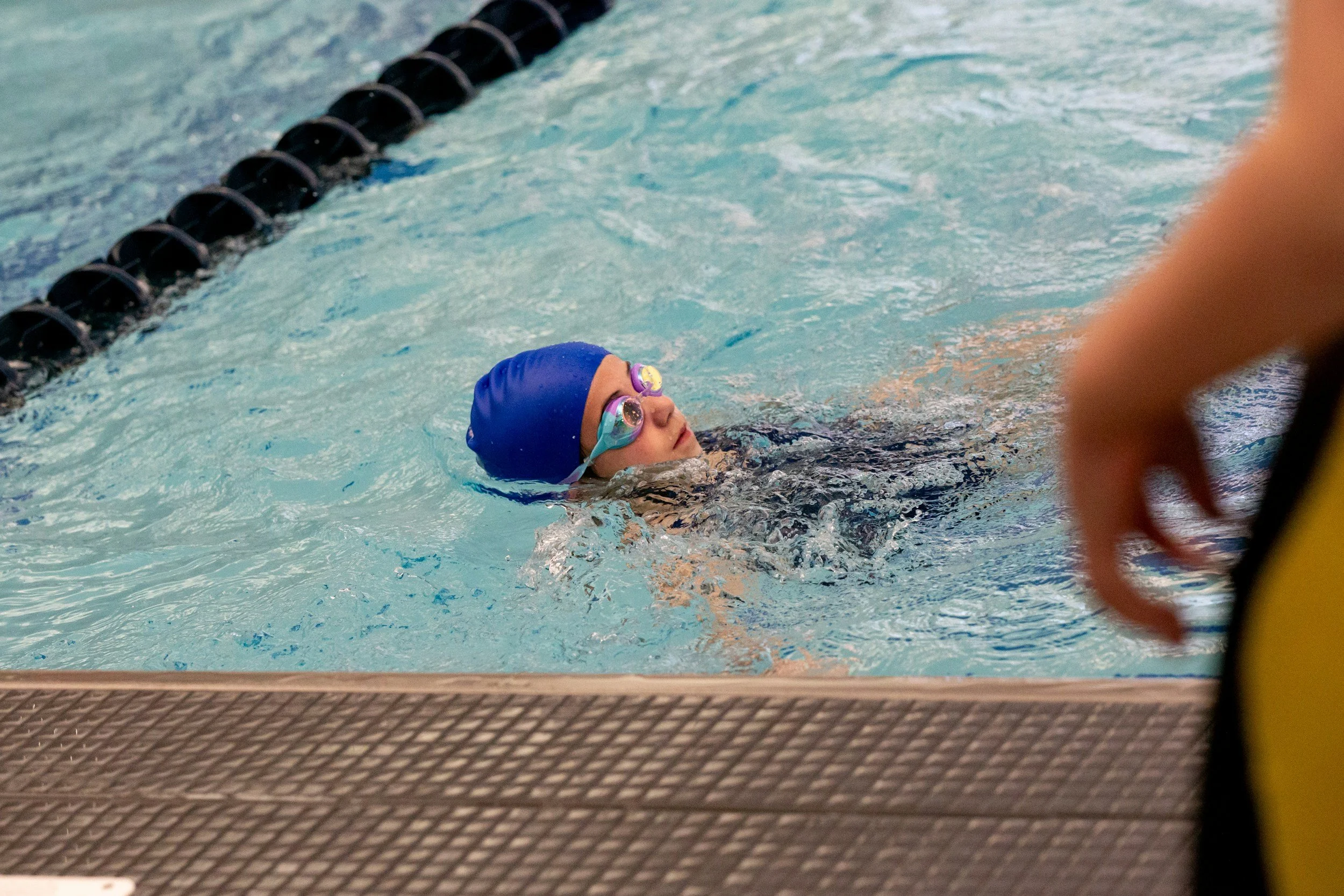 Young swimmer with a blue swim cap and goggles in a pool during a swim meet or practice, with a hand visible in the foreground.
