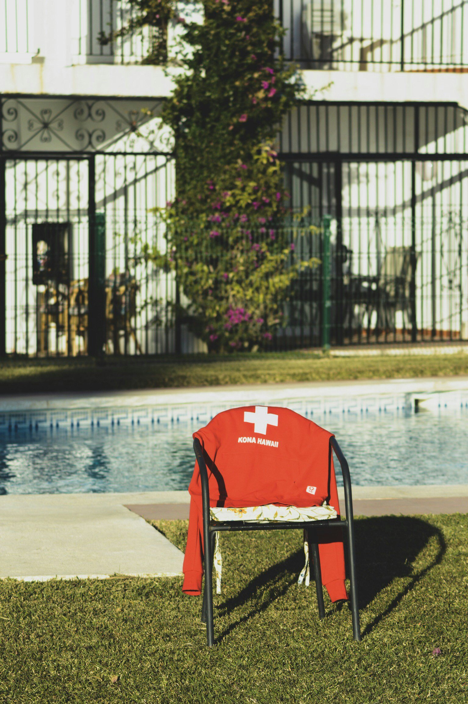 A red lifeguard chair with a white cross and the words 'Kona Hawaii' on the backrest, situated on grass near a swimming pool, with a fenced yard and bushes in the background.