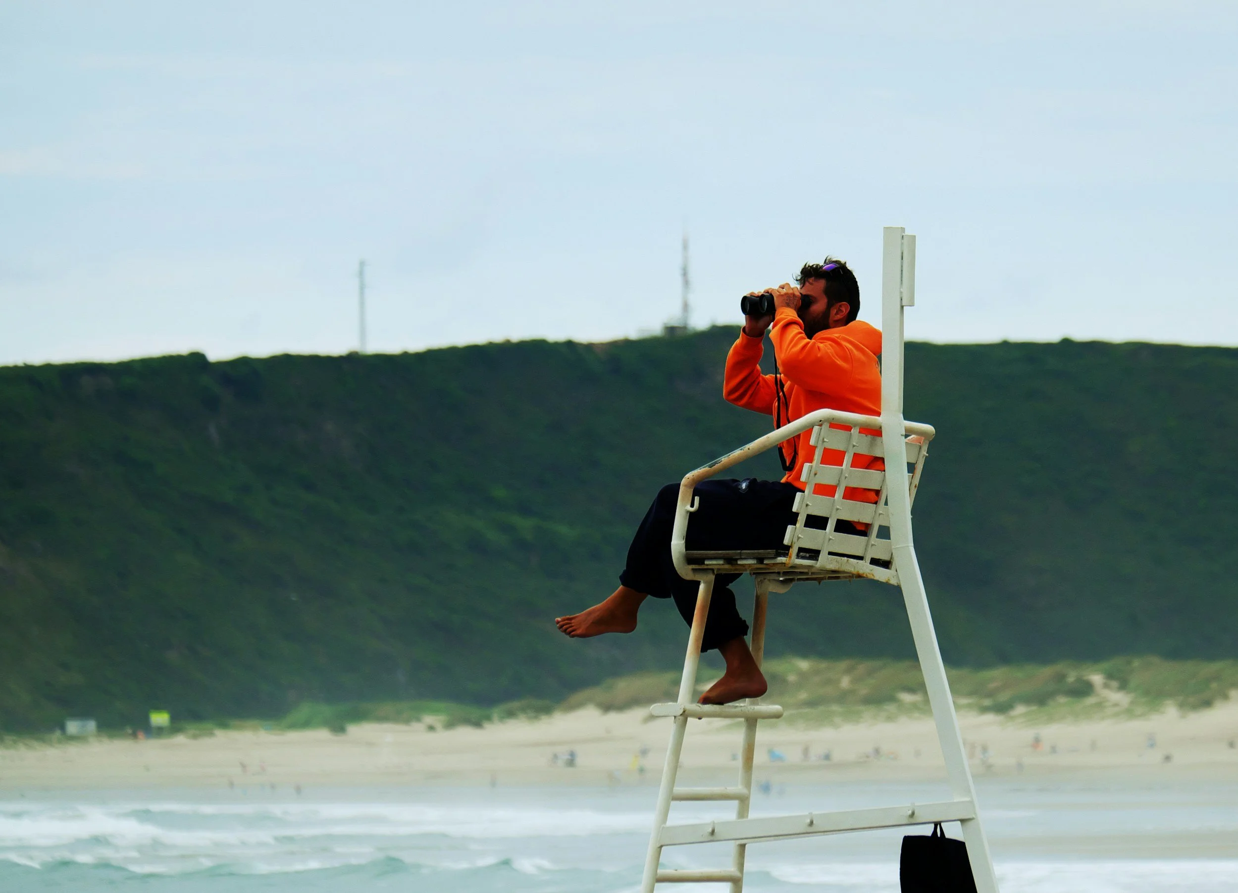 A man sitting on a lifeguard chair on a beach, looking through binoculars at the ocean, with green hills in the background.