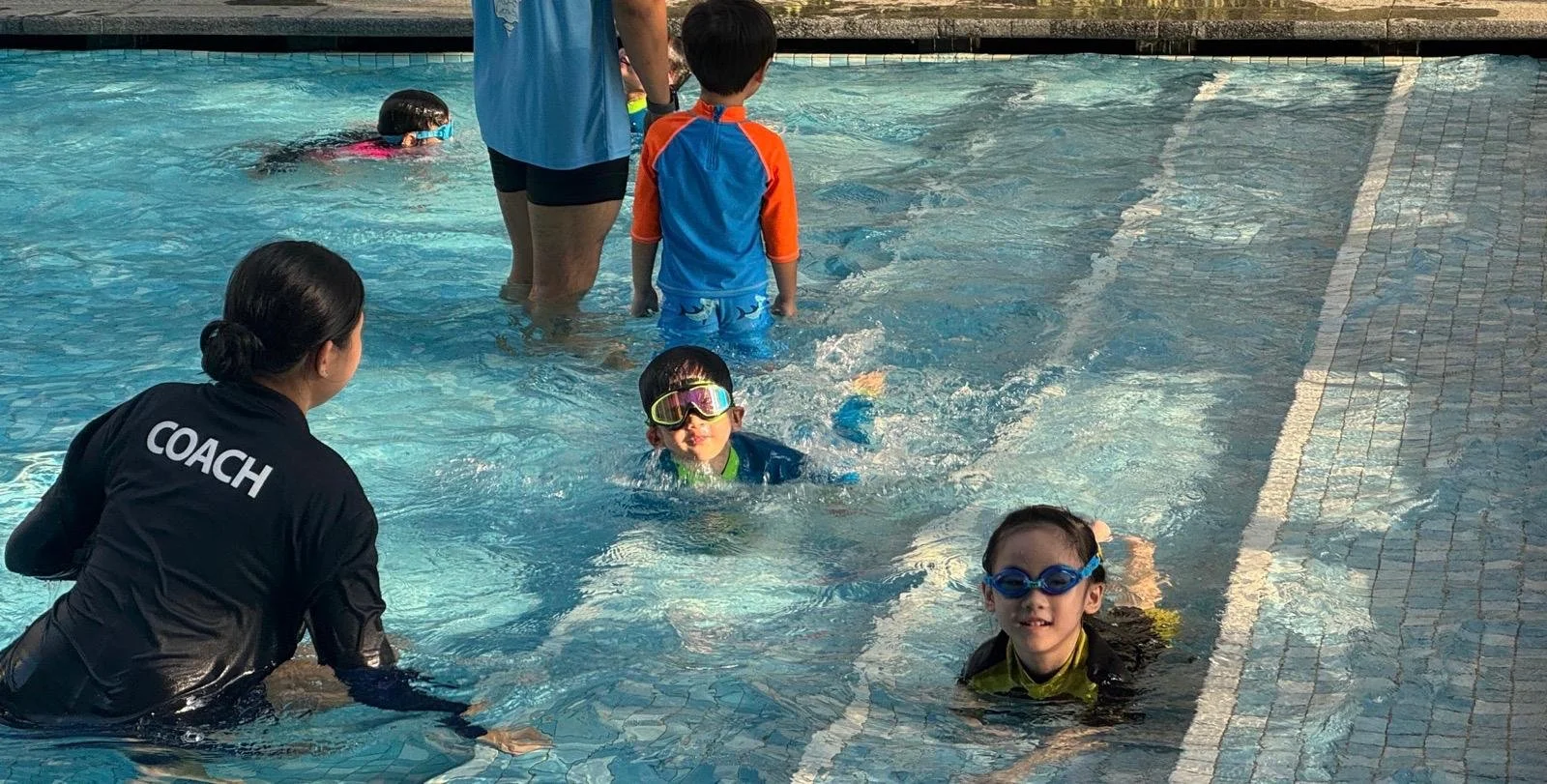 Children learning to swim in a pool with a coach, some kids wearing goggles, and an instructor supervising.