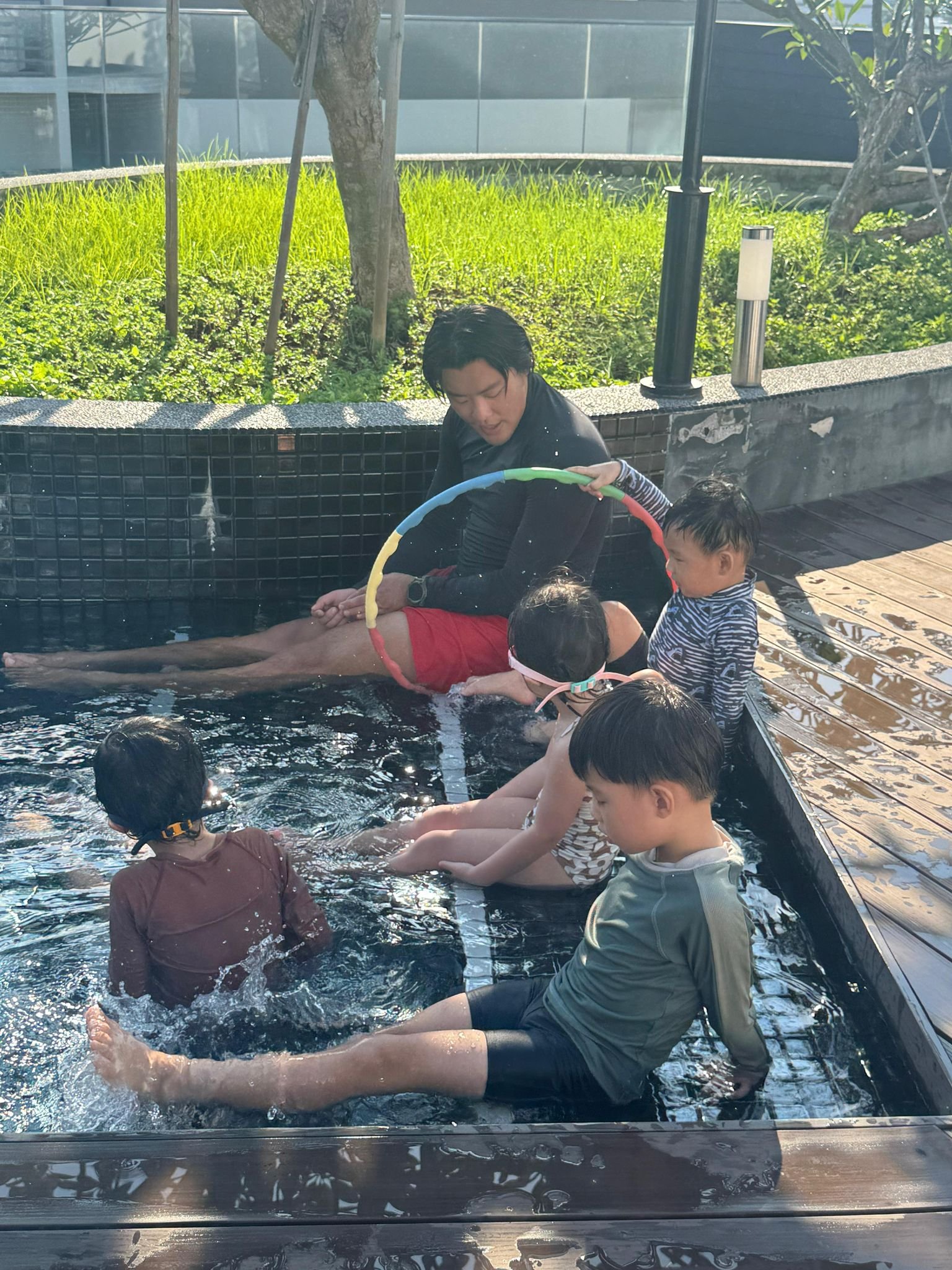 Children sitting in a shallow pool with an adult supervising, playing with a colorful hula hoop outdoors on a sunny day.
