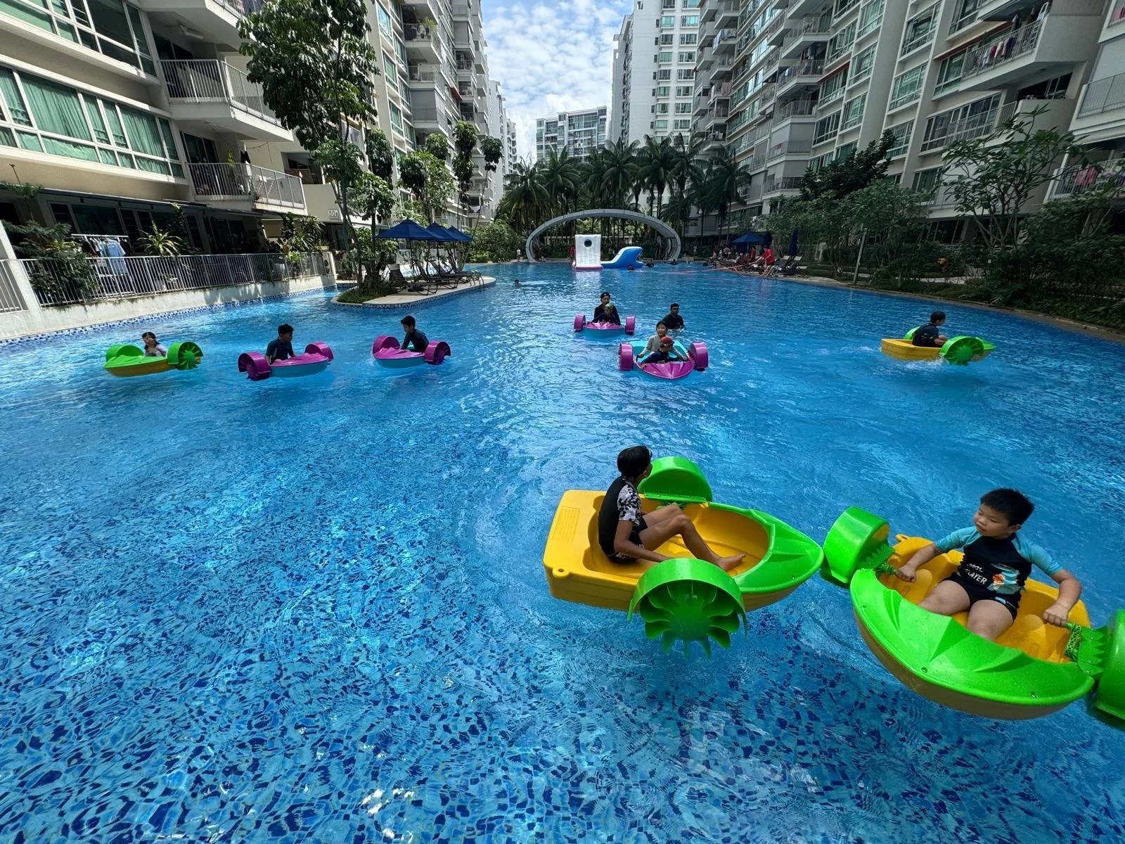 Children riding small colorful pedal boats on a swimming pool surrounded by high-rise apartment buildings with trees and lounge chairs.