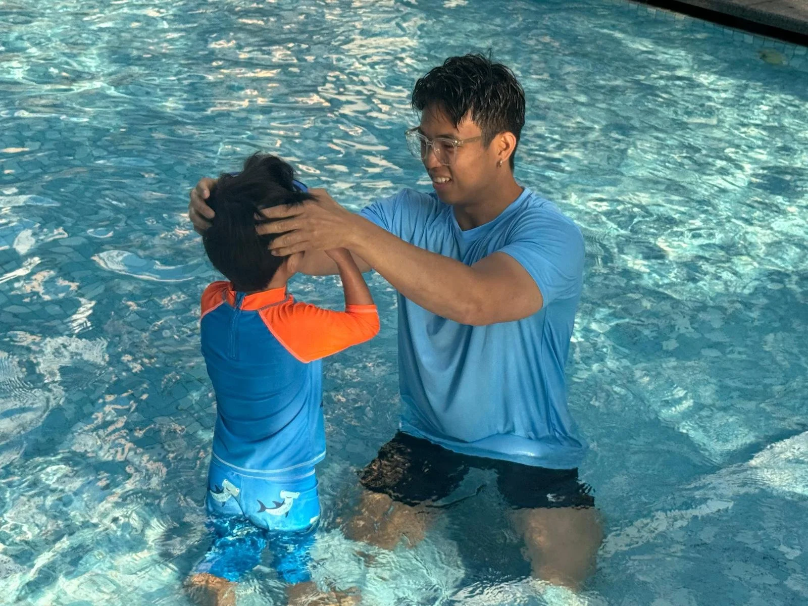 A man in a blue t-shirt is helping a young boy in a blue and orange swimming shirt and dolphin swim trunks in a pool. The man is holding the boy's head and face in a teaching or guiding manner, with water around them.